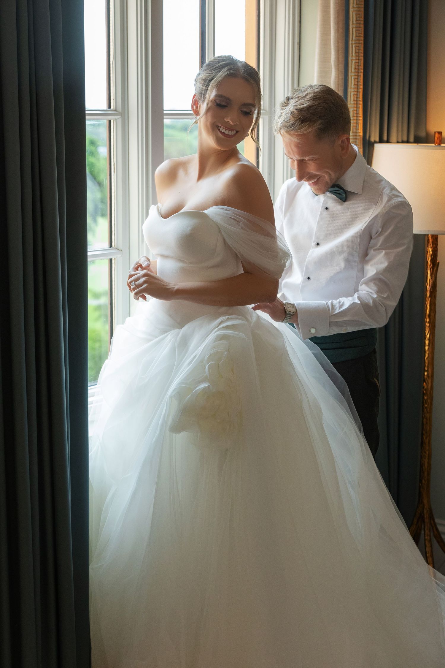 Bride in off-shoulder white tulle ball gown smiles as groom in white shirt fastens her dress by a sunlit window.