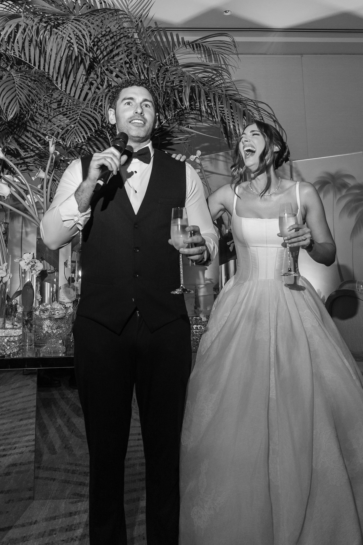 Black and white photo of a groom speaking into a microphone and bride laughing at their wedding reception.