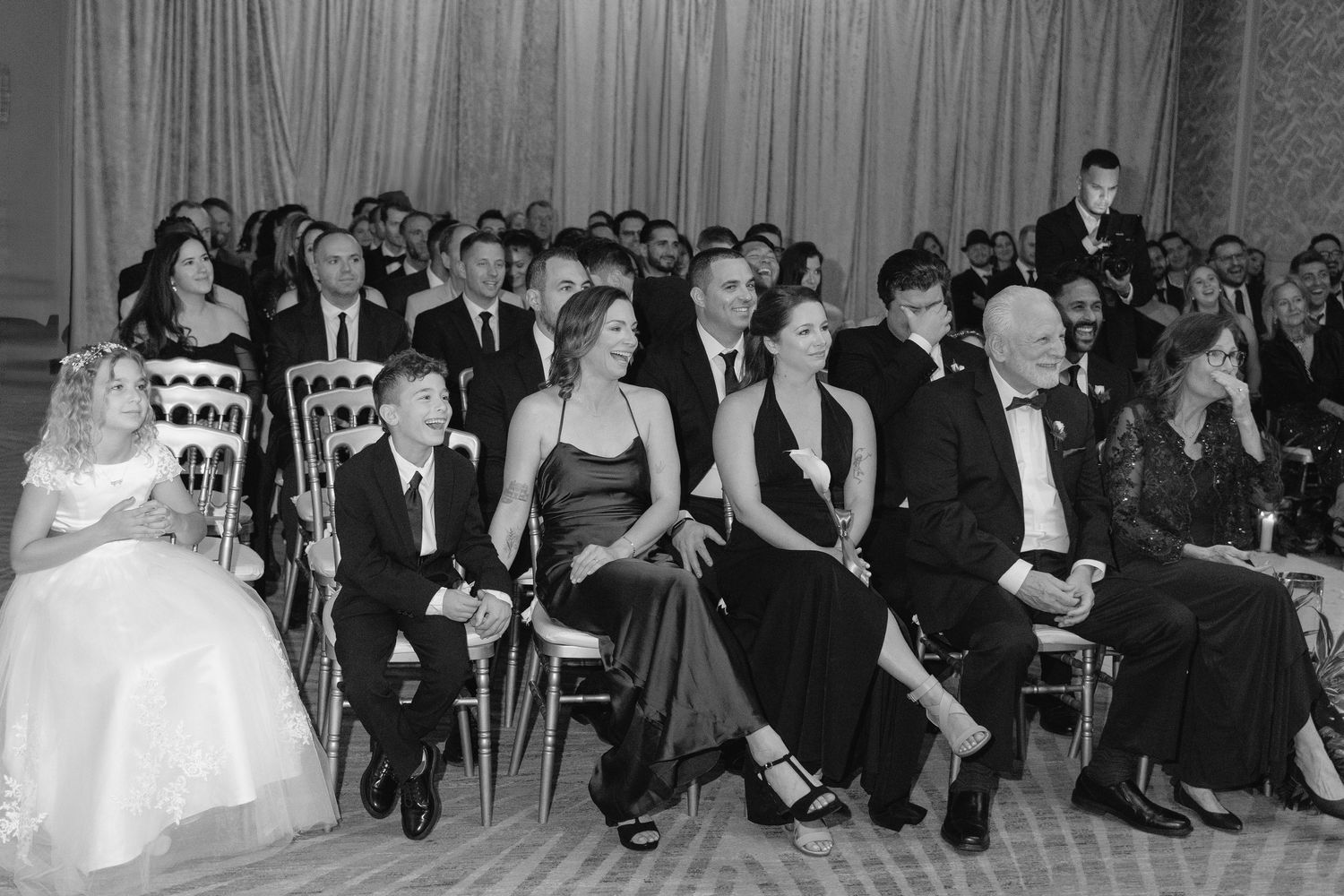 Black and white photo of elegantly dressed wedding guests seated and smiling during a formal indoor ceremony.