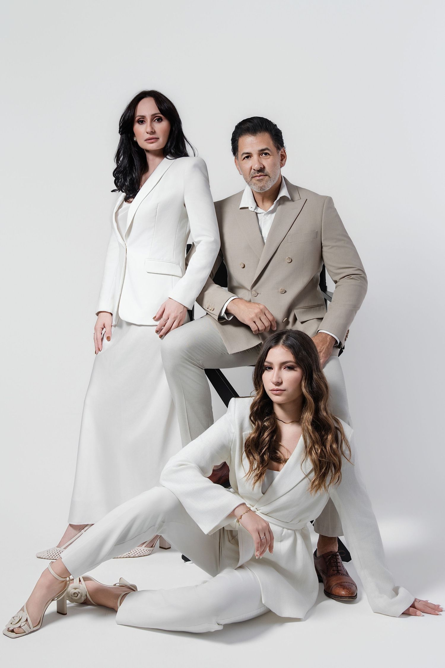 Three elegantly dressed people pose in white and beige outfits against a white background in a professional photo.