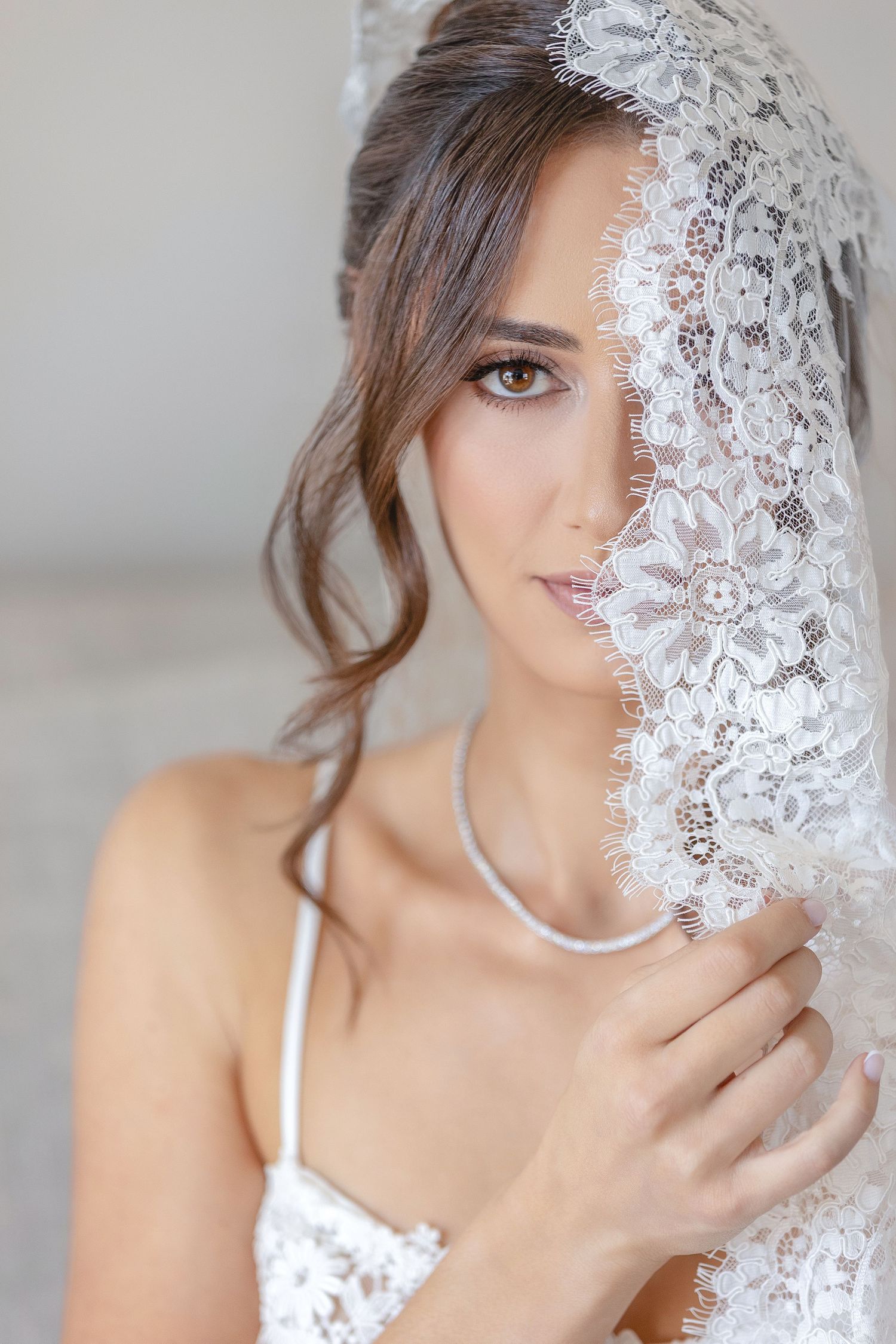 Elegant bride holding white lace veil over face, wearing diamond necklace and lace dress, soft studio lighting.