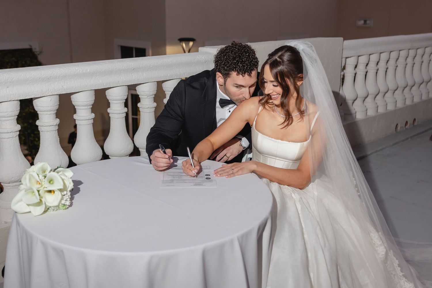 Bride and groom signing wedding documents at a white table, with a bouquet of white flowers nearby.