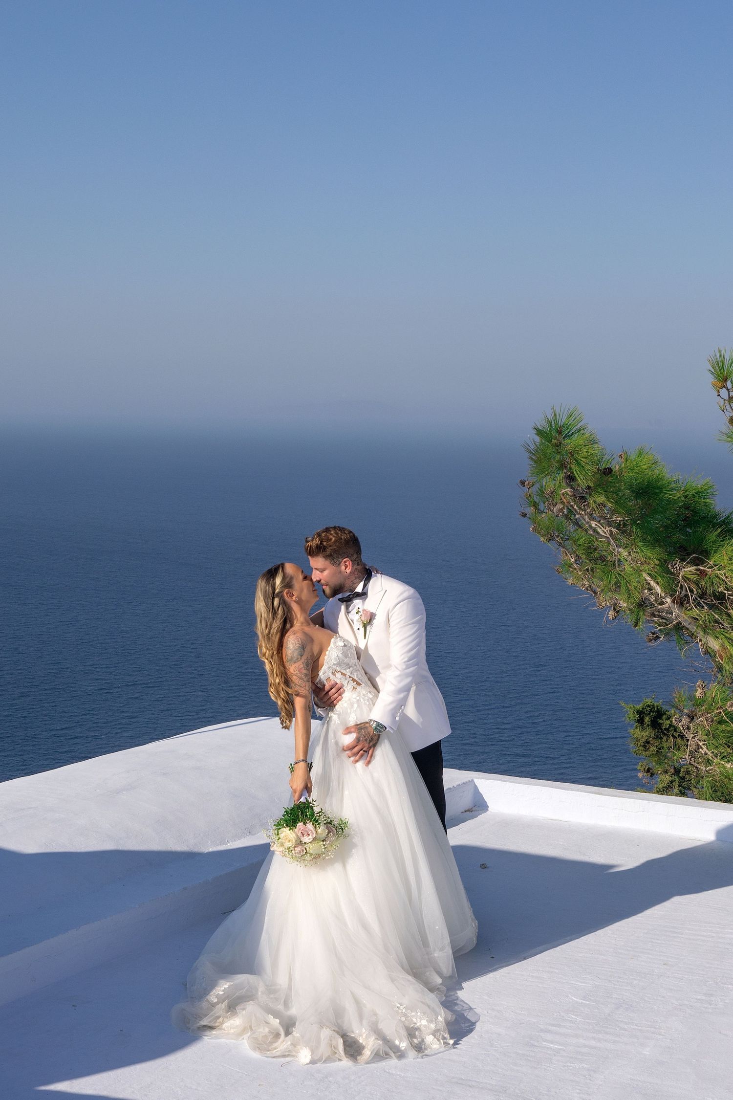 Bride and groom kissing on white terrace overlooking blue sea, bride in white gown holding bouquet, groom in white tuxedo.