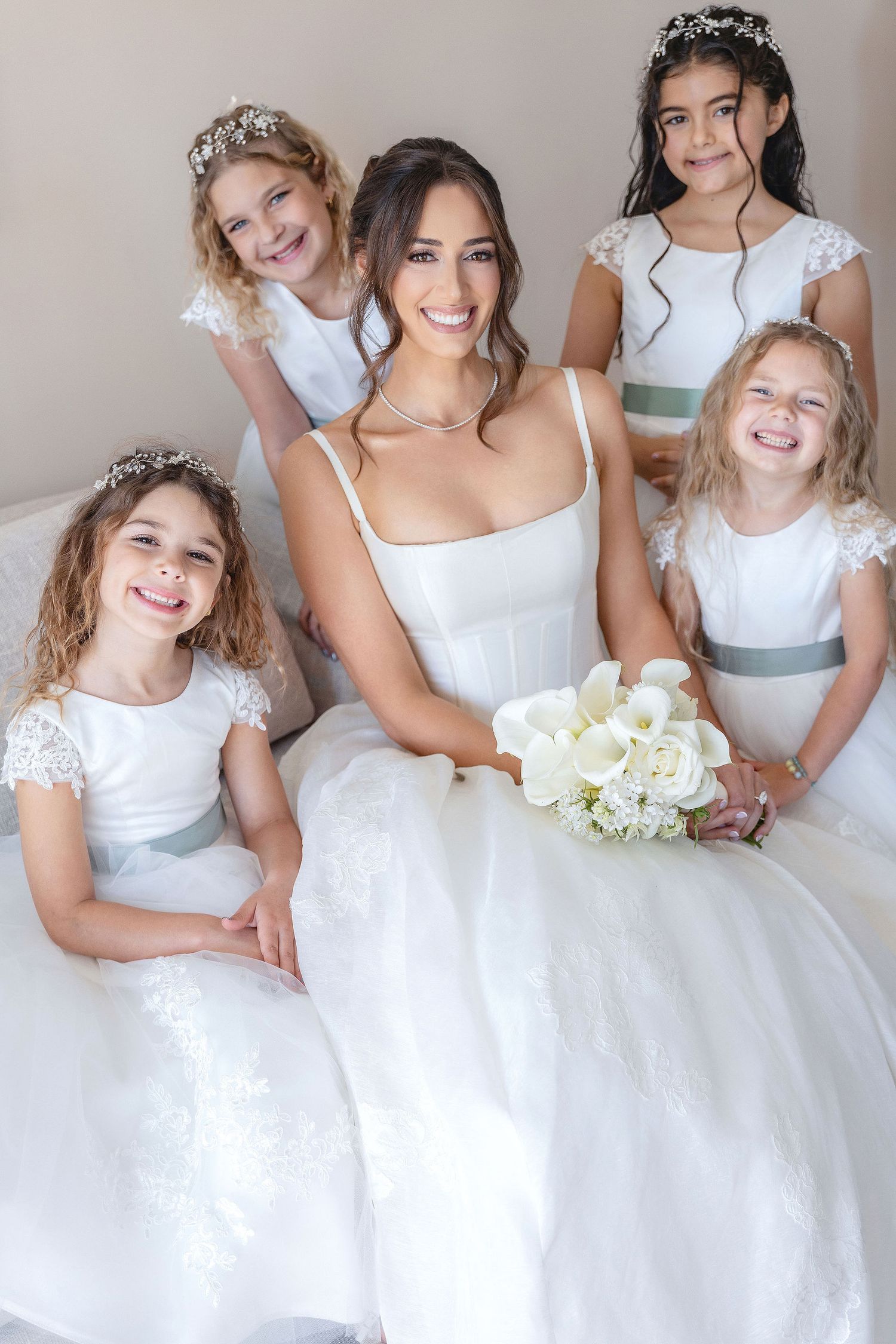 A bride in a white gown poses with flower girls wearing matching white and gray dresses and holding a white bouquet.