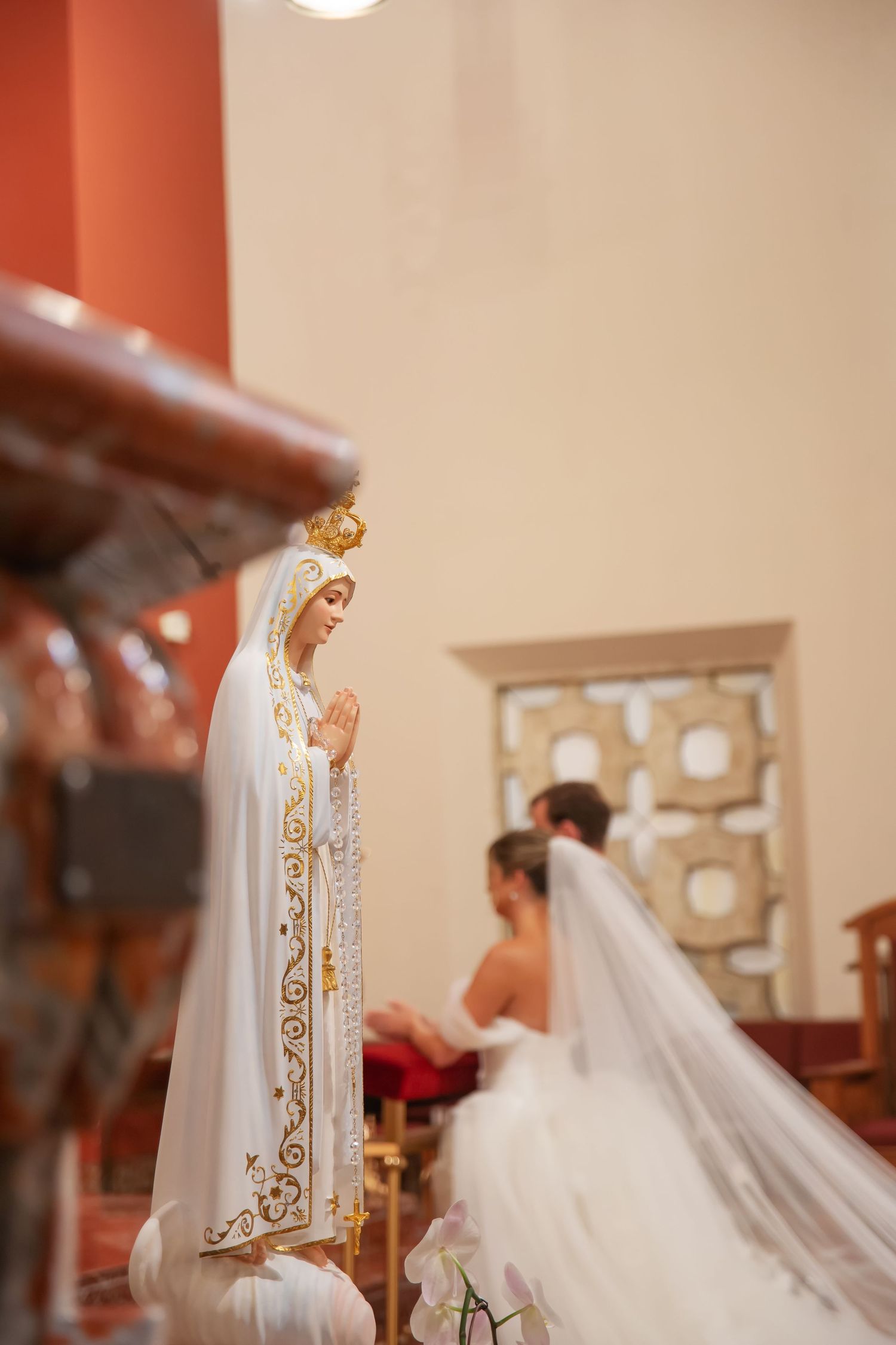 Catholic wedding ceremony with bride in white gown and veil kneeling at altar beside Our Lady of Fatima statue.