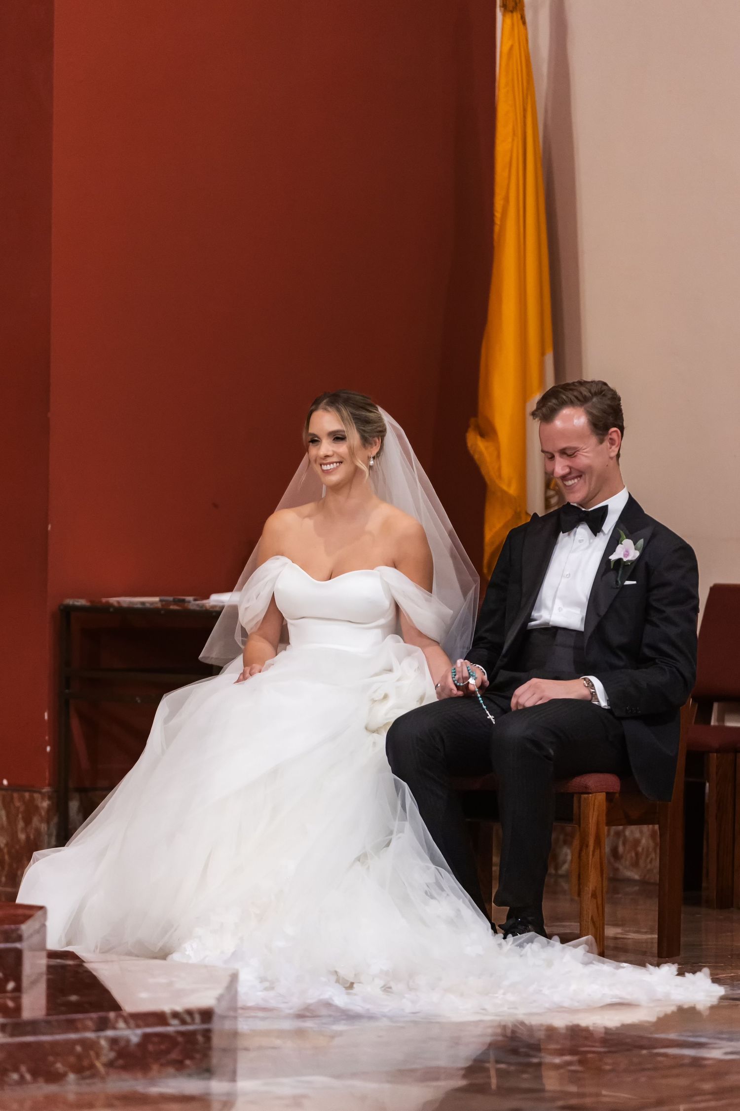 Bride in white off-shoulder gown and veil sits beside groom in black tuxedo during wedding ceremony.