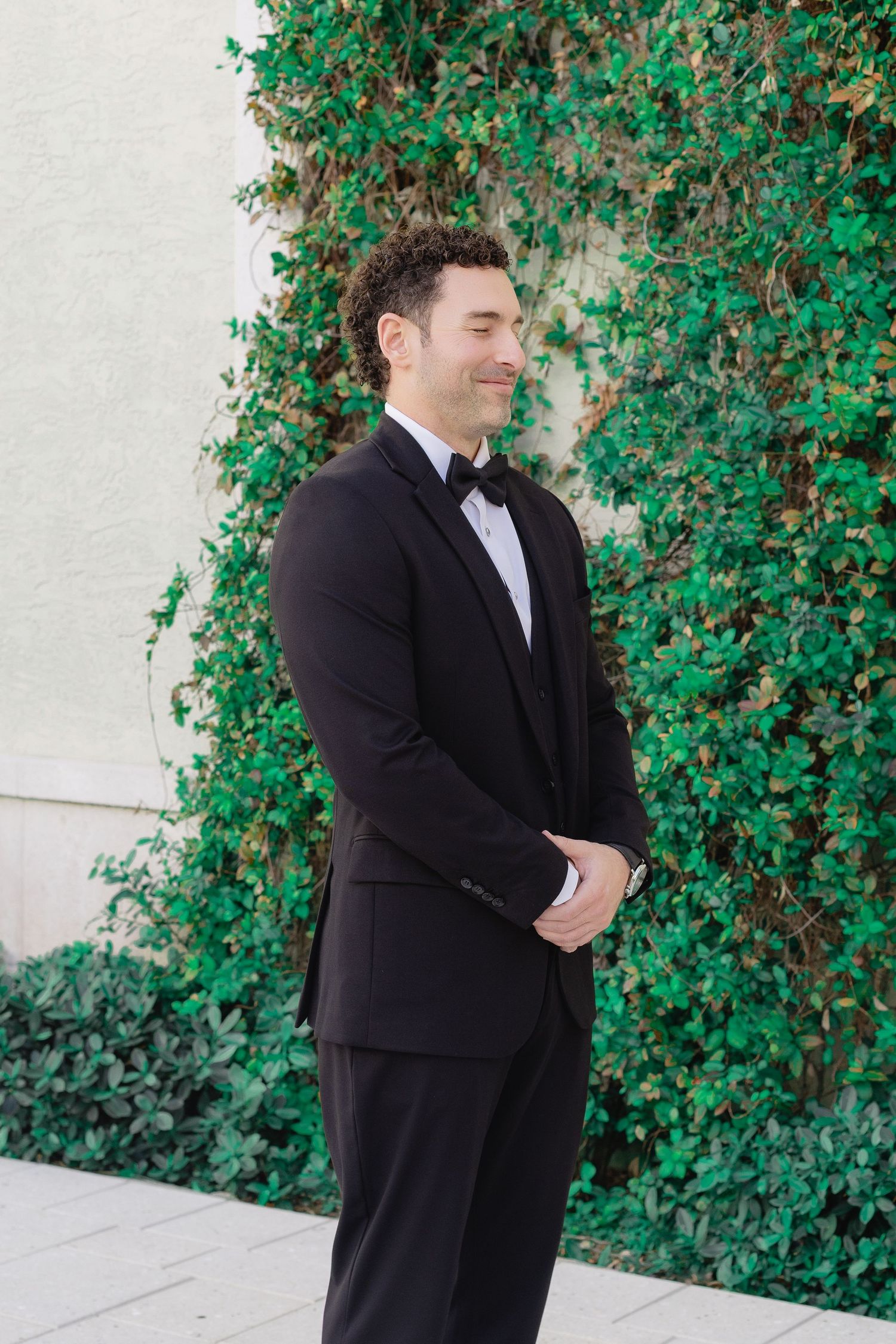 A groom in a black tuxedo with bow tie smiles while standing in front of a lush green ivy-covered wall.