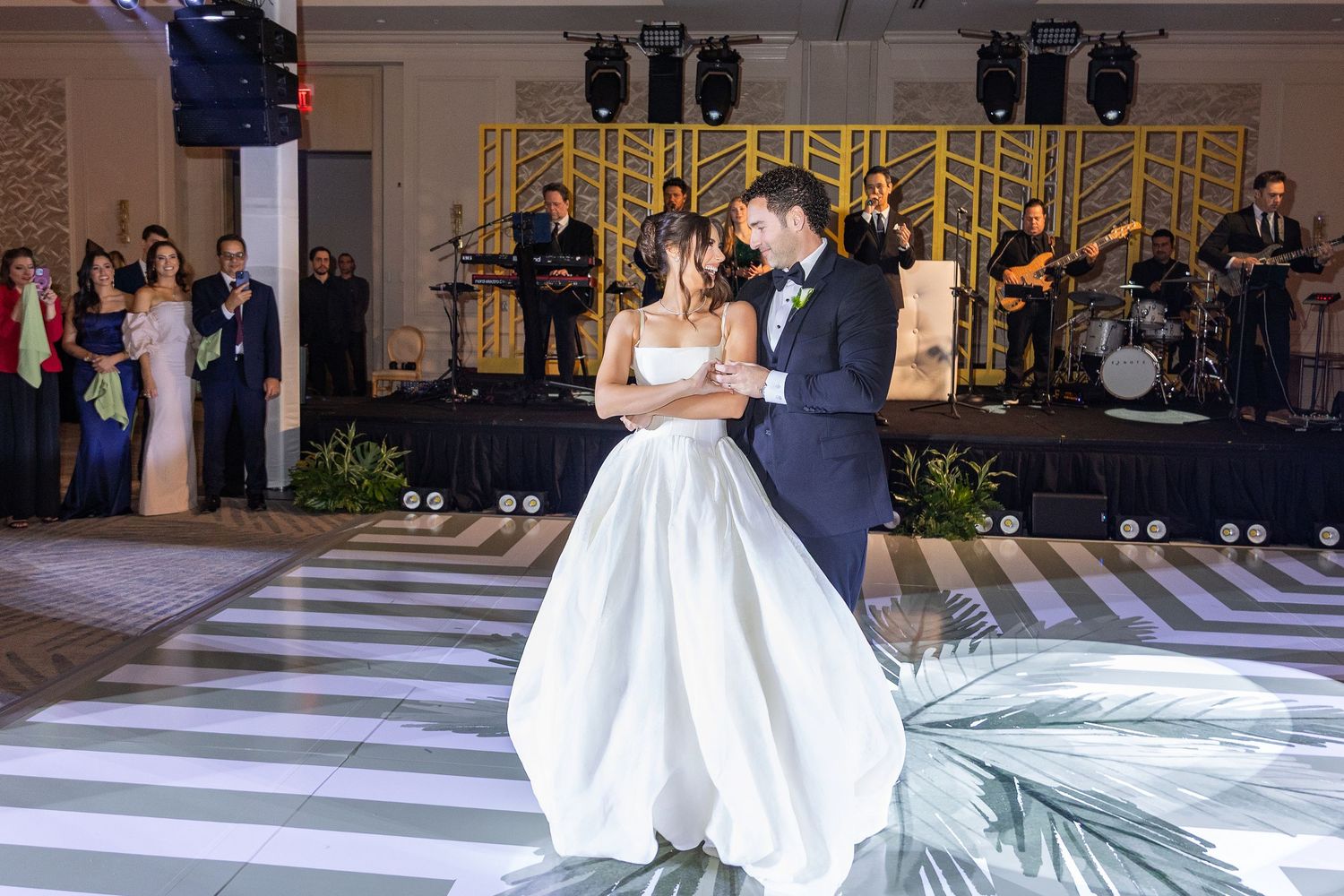 Bride and groom share first dance at elegant wedding reception with live band performing on stage in background.