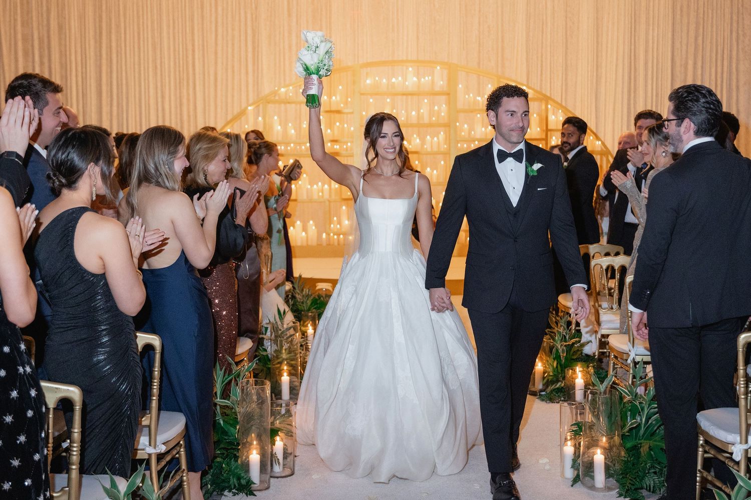 Bride and groom walk down aisle after wedding ceremony, bride raising bouquet, guests celebrating in elegant venue.