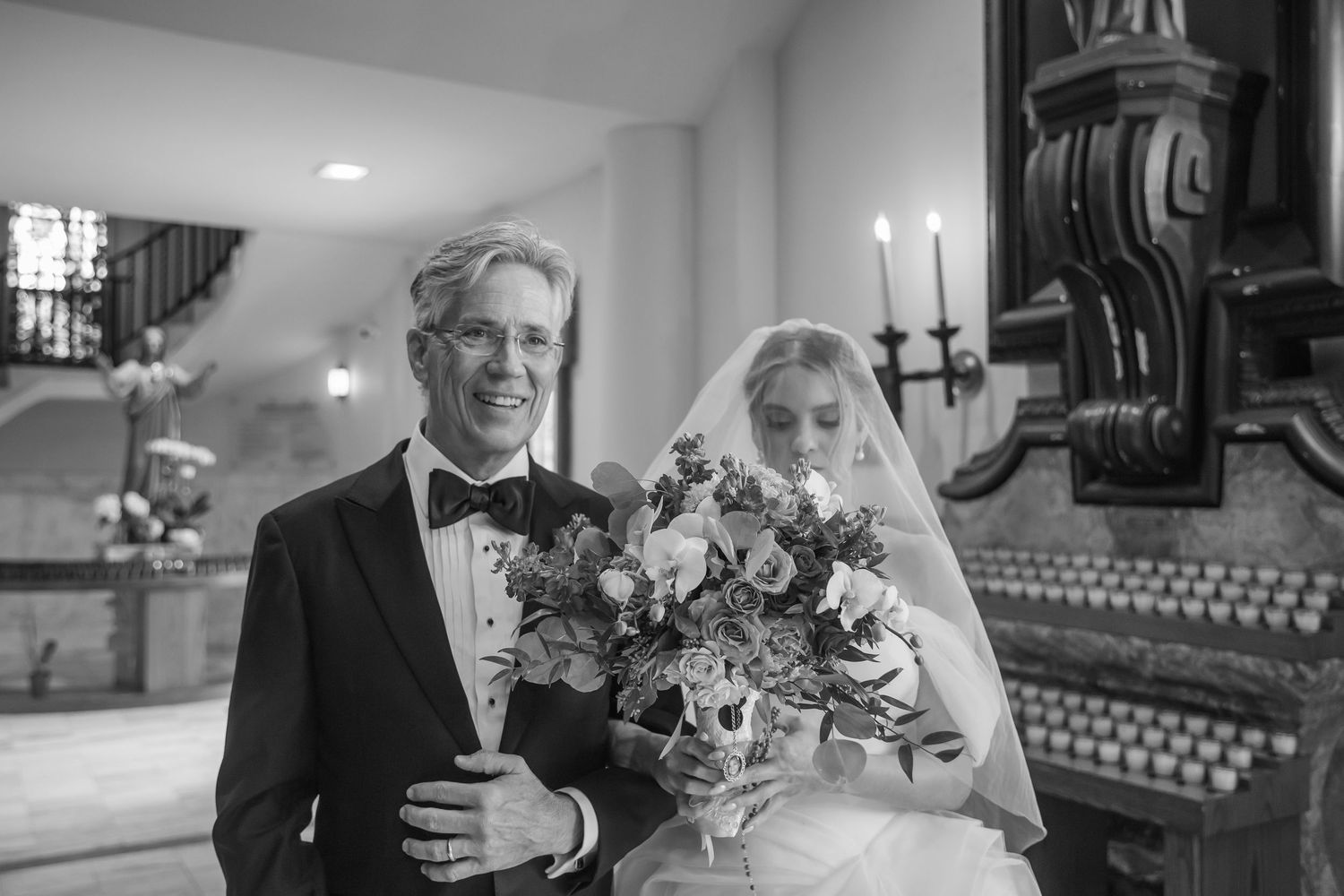 Black and white photo of a smiling father in a tuxedo walking his bride daughter down the aisle.