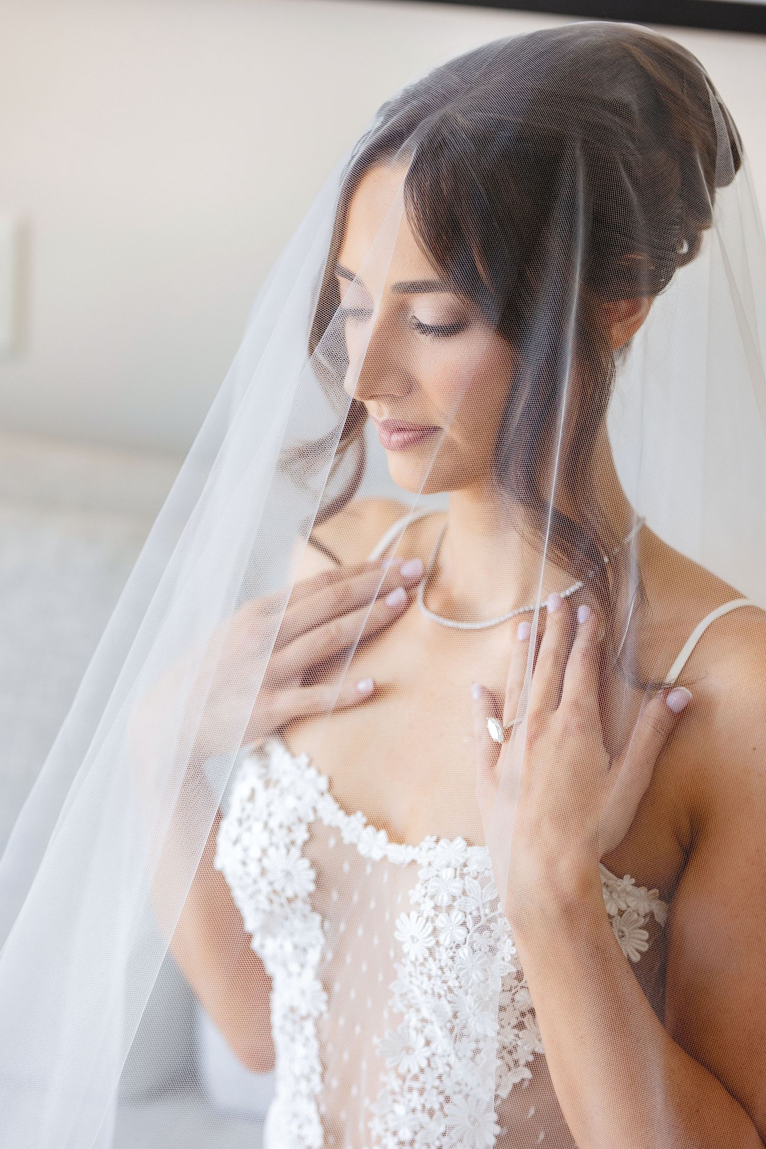 Elegant bride in lace wedding dress with sheer veil draped over her, touching necklace with eyes closed.