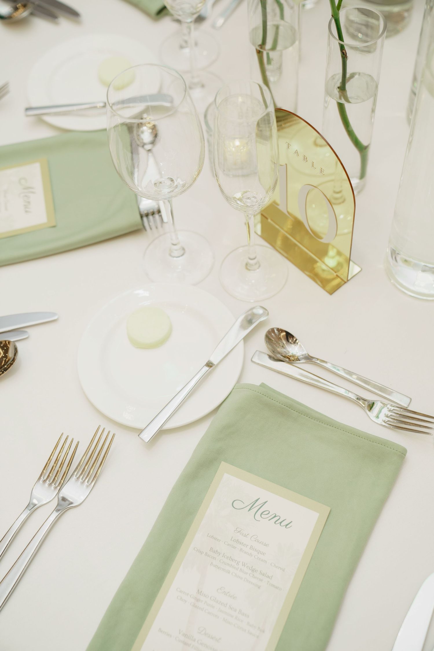 Elegant wedding table setting with sage green napkins, gold table number, crystal glasses, and silver cutlery on white tablecloth.