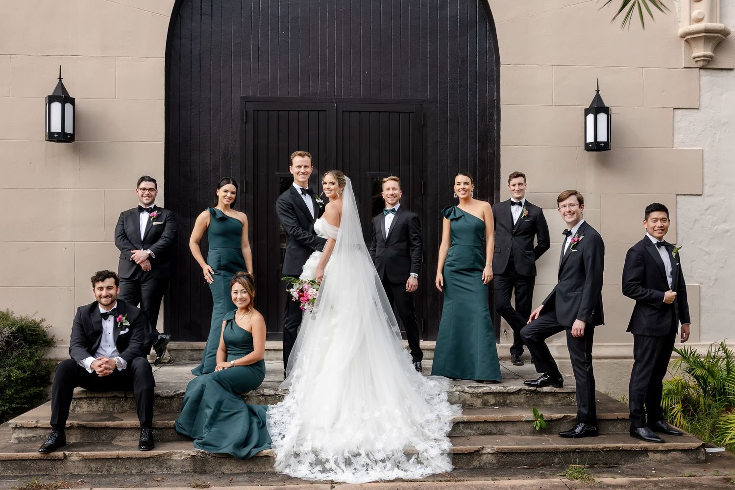 Wedding party poses on steps, bride in white gown with groomsmen in black tuxedos and bridesmaids in emerald green gowns.