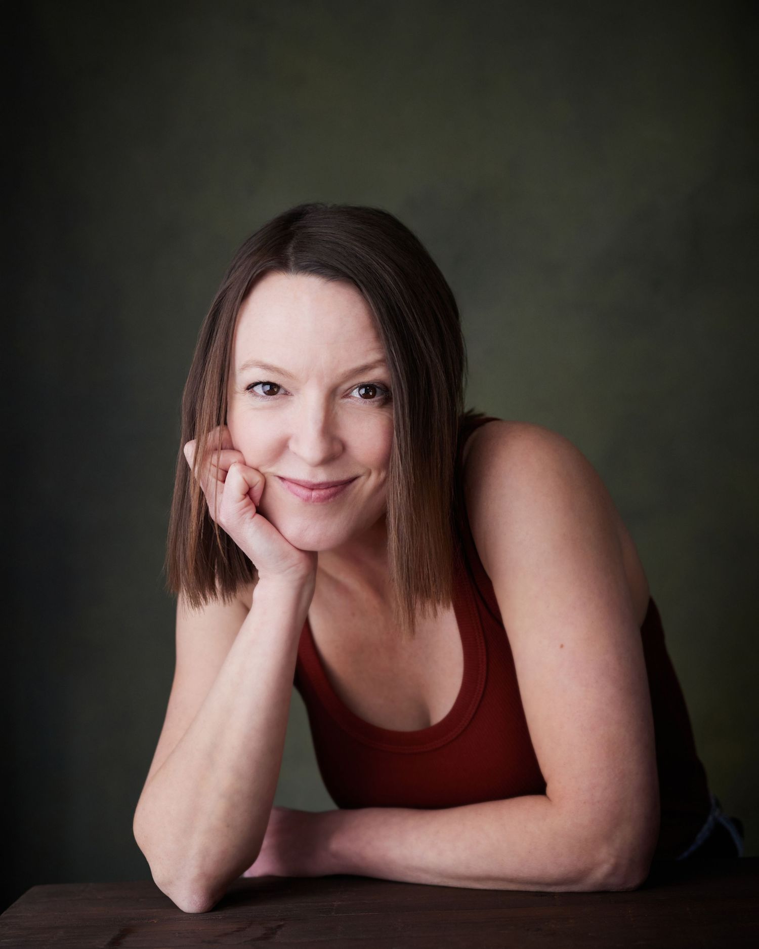 Relaxed headshot; actress with a welcoming smile leaning on her hand against a green background.
