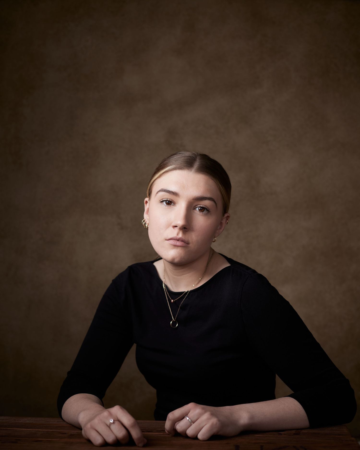 Edgy actor headshot; woman seated at a table with a cool, detached gaze on brown.