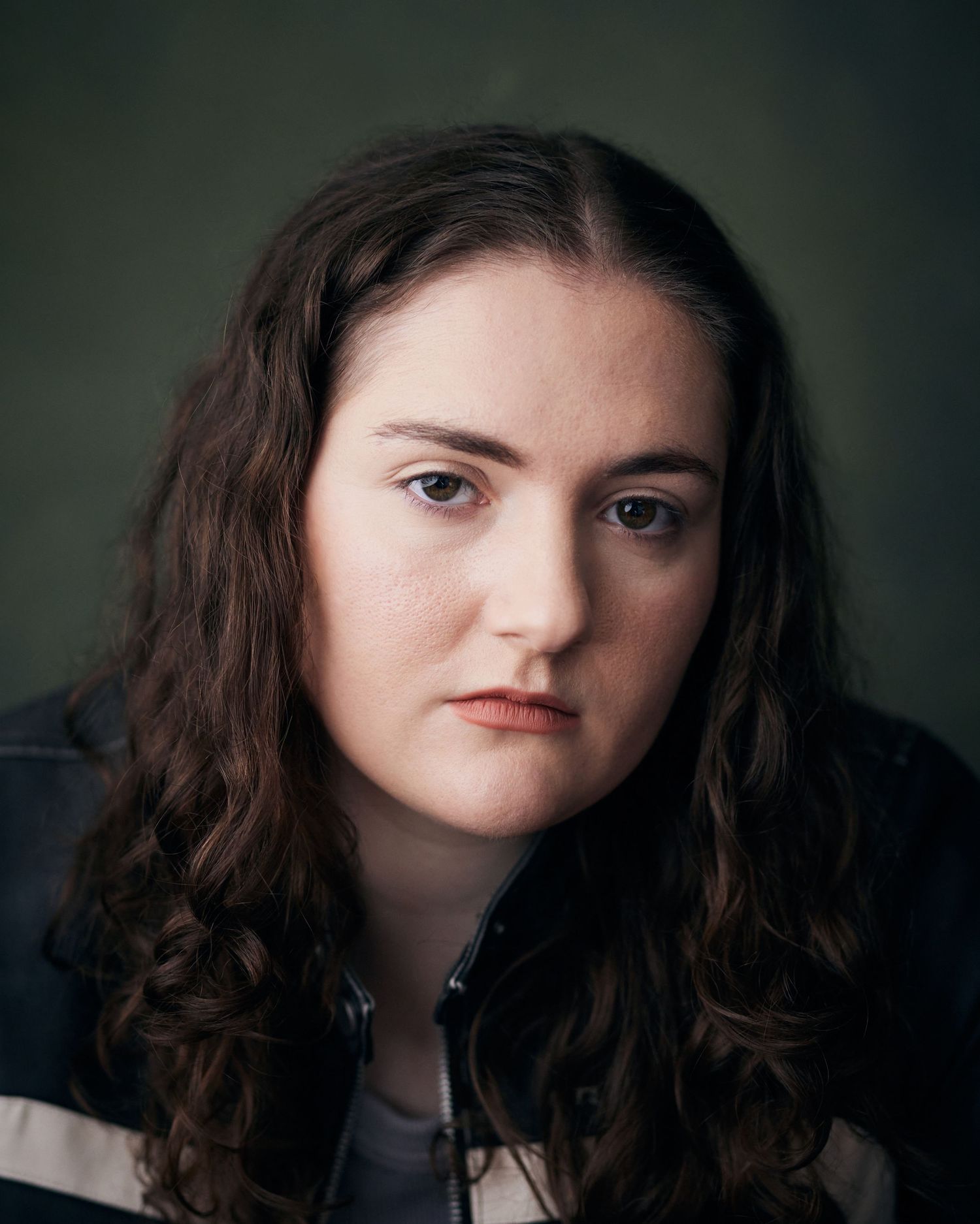 Dramatic actor headshot; woman with a piercing, serious gaze against a dark green textured backdrop.