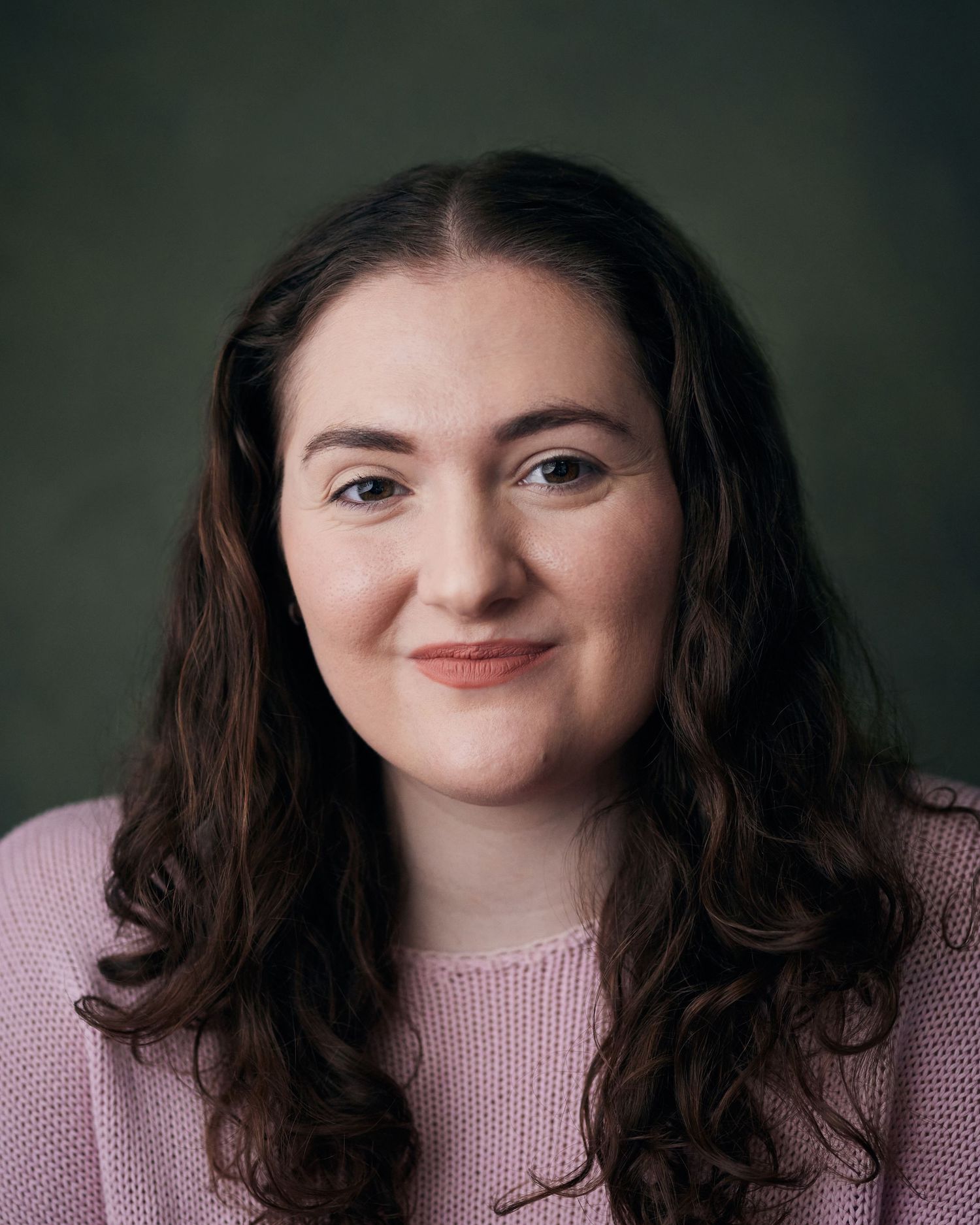 Relaxed headshot; actress with natural wavy hair and a bright, welcoming smile against a sage green background.