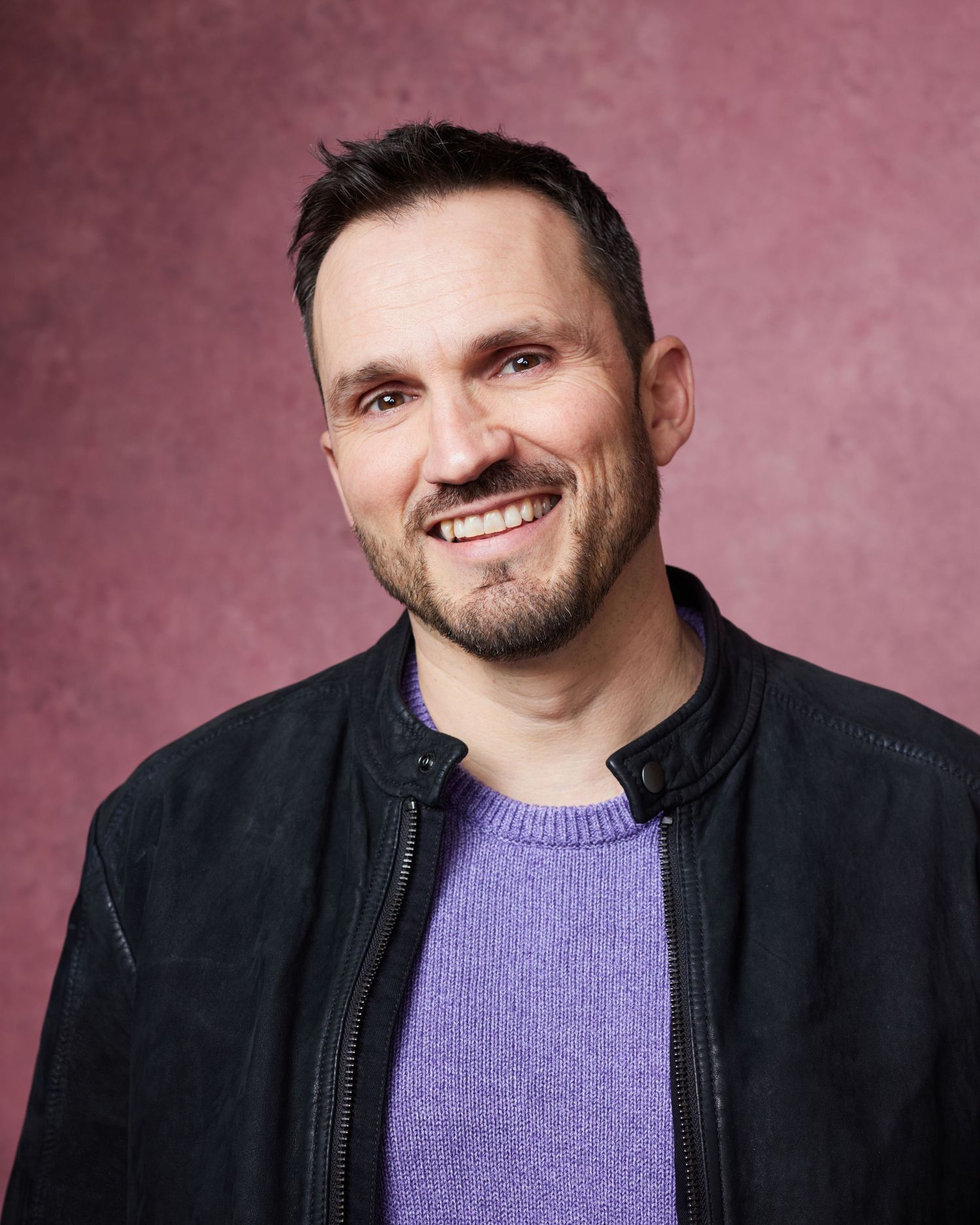 Cheerful studio headshot; man with a bright, genuine, and wide-eyed smile against a pink canvas backdrop.