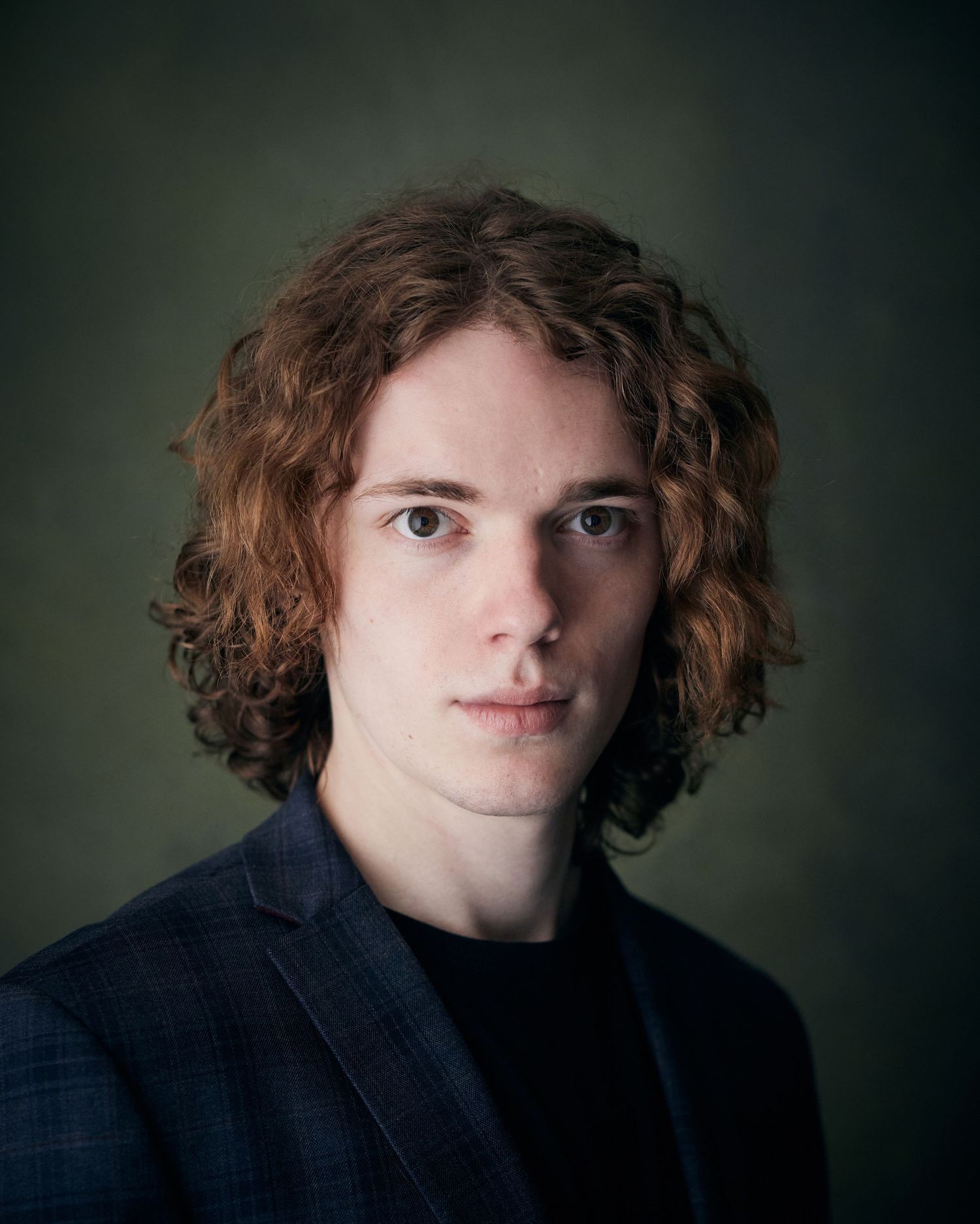 Professional Cardiff actor headshot of a man with curly hair wearing a dark blazer and black tee against a textured green background.