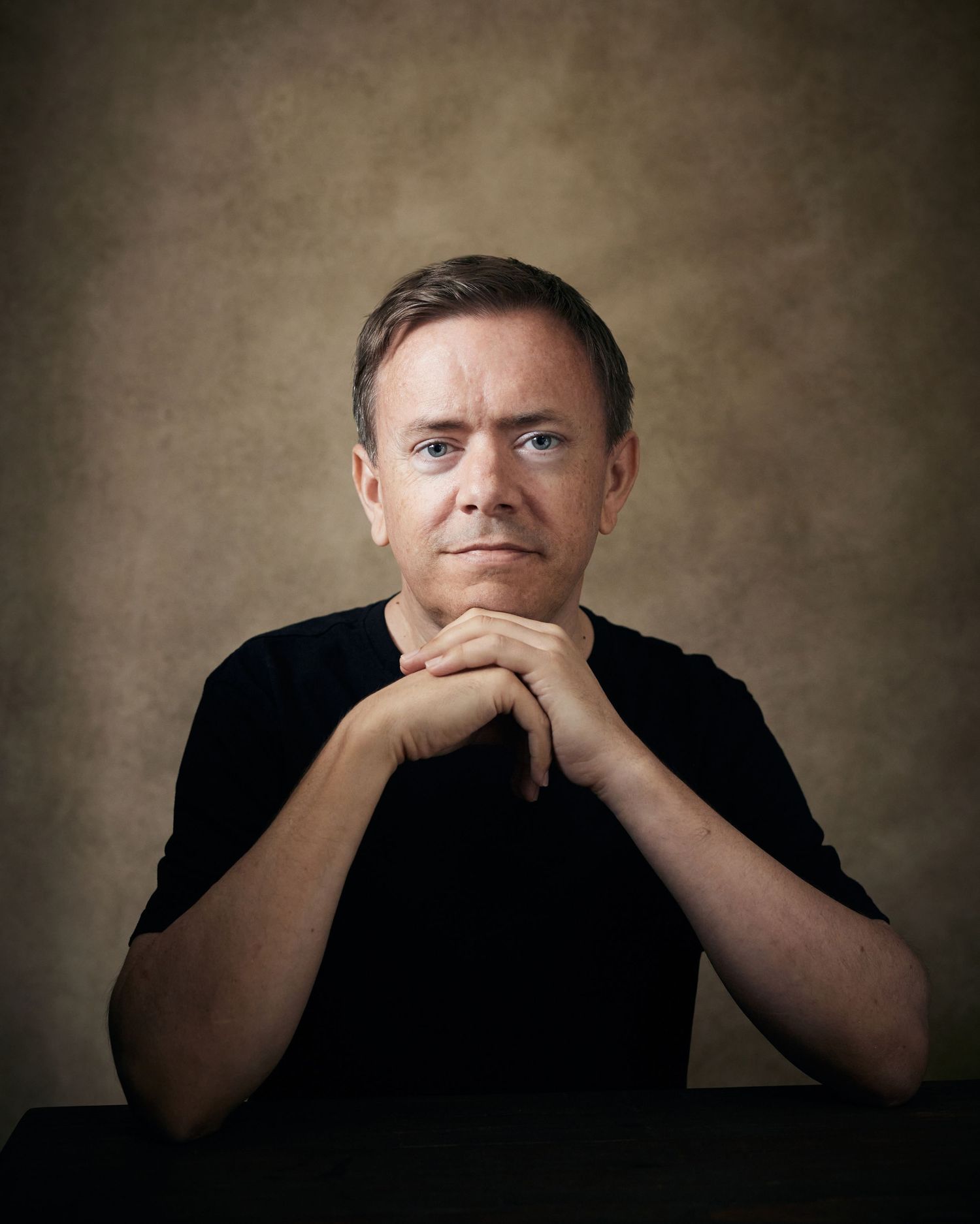 Approachable character headshot; male performer with a bright, open presence, seated against a brown canvas backdrop.