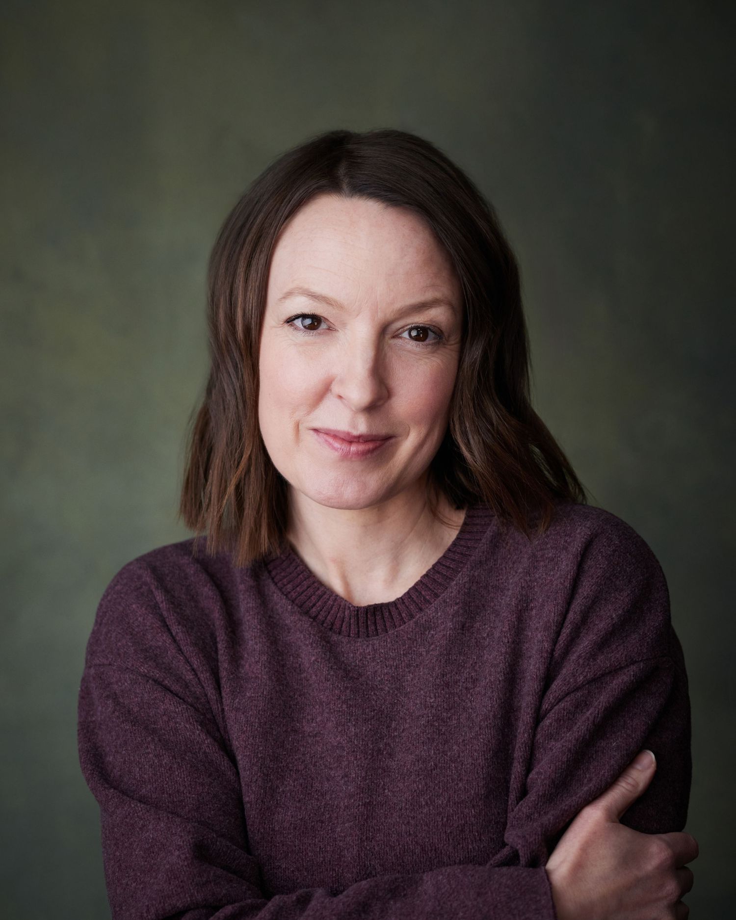 A warm studio portrait of a woman in a purple jumper against a green canvas background.