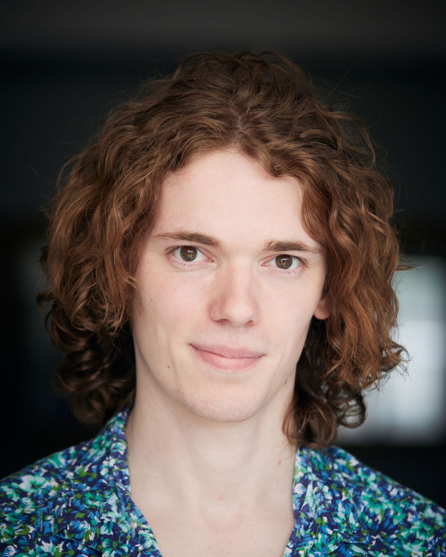 Warm, approachable actor headshot of a man with curly hair wearing a blue floral shirt against a soft, bright background in Cardiff.