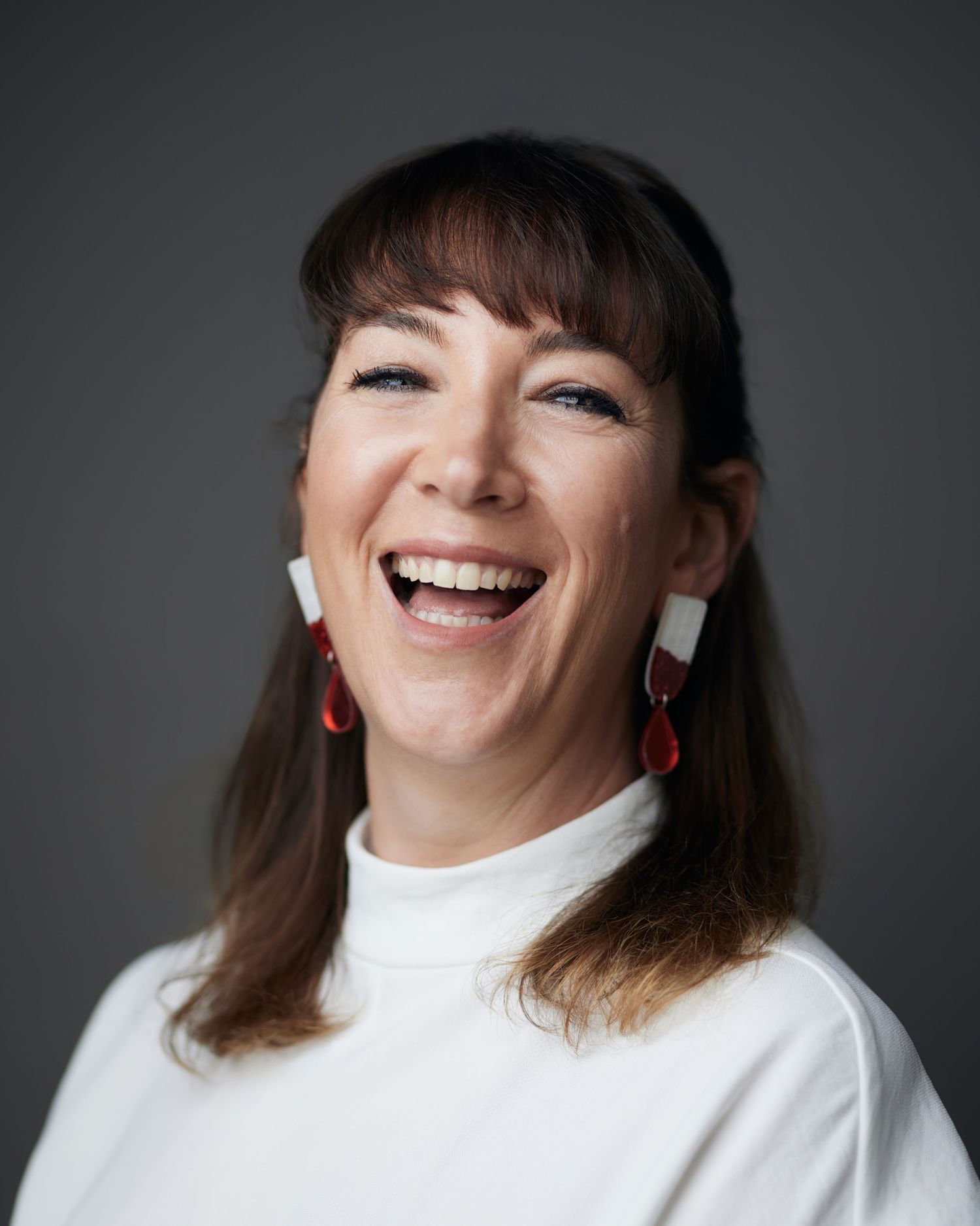 Friendly business portrait by Geraint Todd; woman with a joyful, open laugh and brown hair with a fringe, wearing bold red-accented earrings