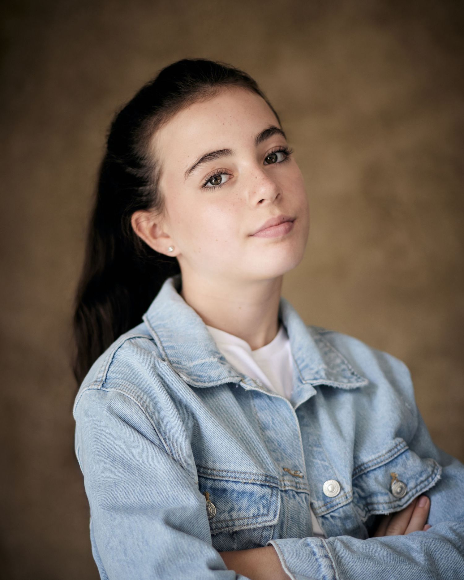 Edgy child actor headshot; girl in a denim jacket with a cool, confident gaze on brown.