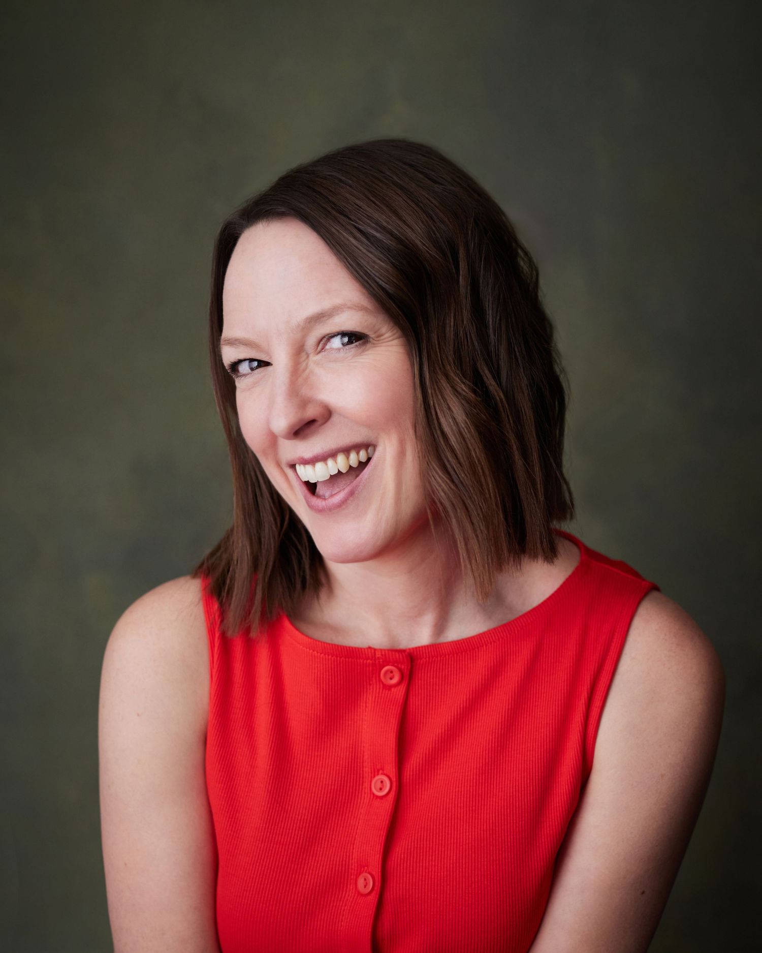 Commercial actor headshot; woman with a big smile in a red top against a green canvas backdrop.