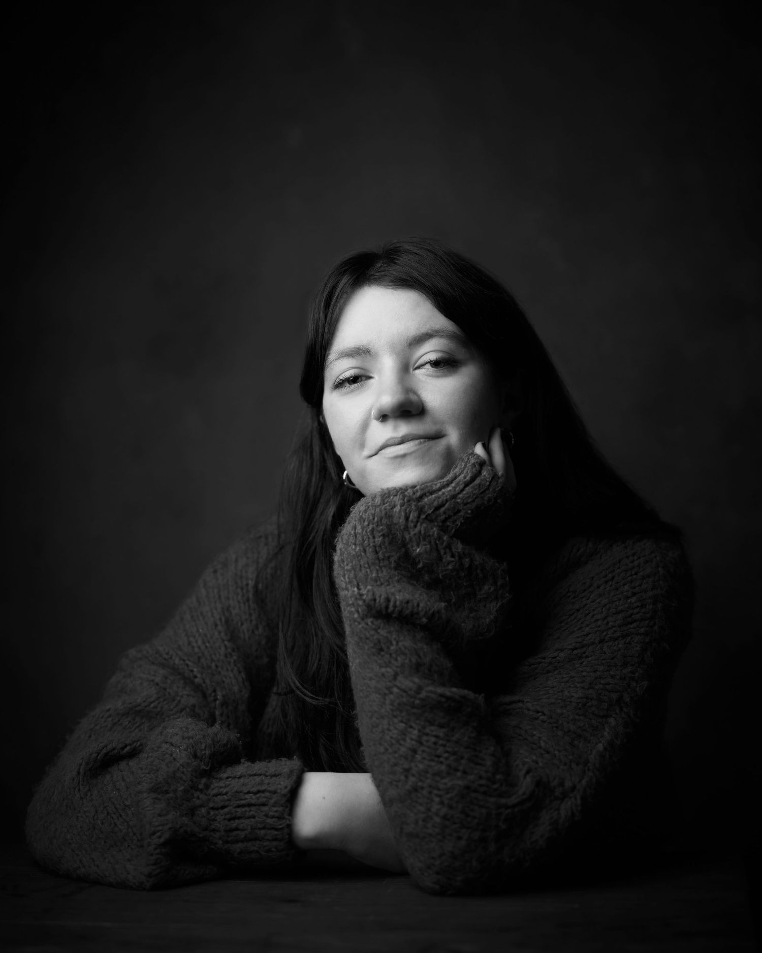 Confident actor headshot: a black and white photo of a woman smiling confidently, seated a table and leaning on her hand.