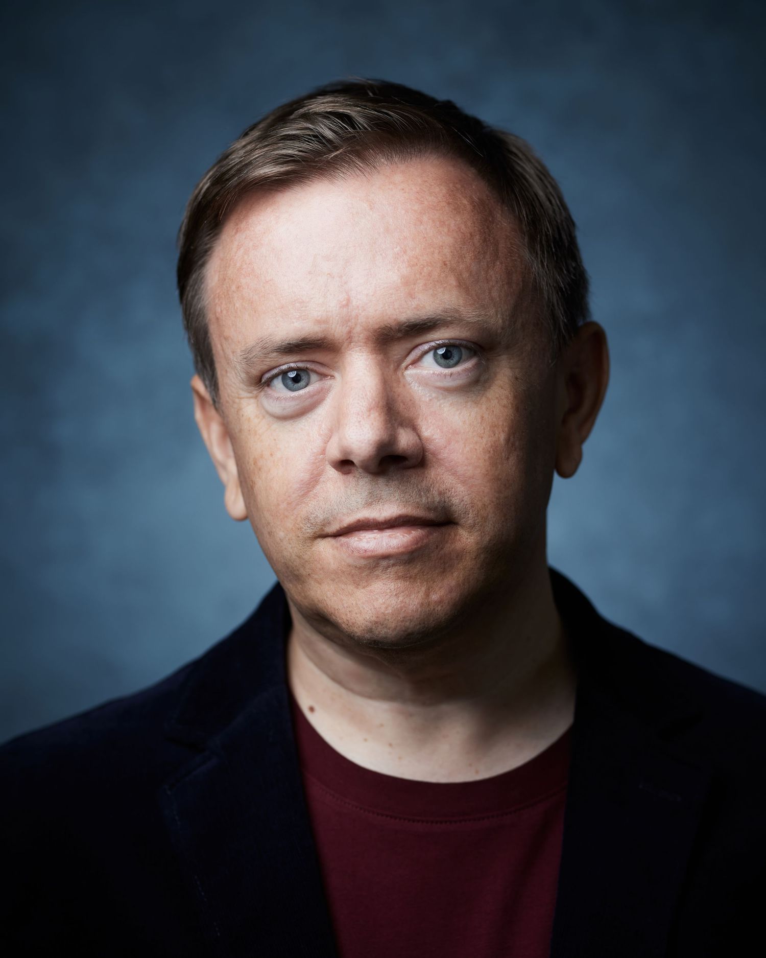 Professional neutral headshot; male performer with a calm, steady gaze against a blue background.