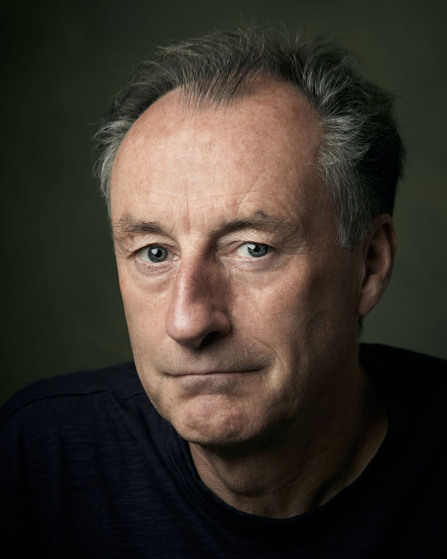 Professional neutral headshot; male performer with a calm, steady  and confident gaze against a green canvas backdrop.