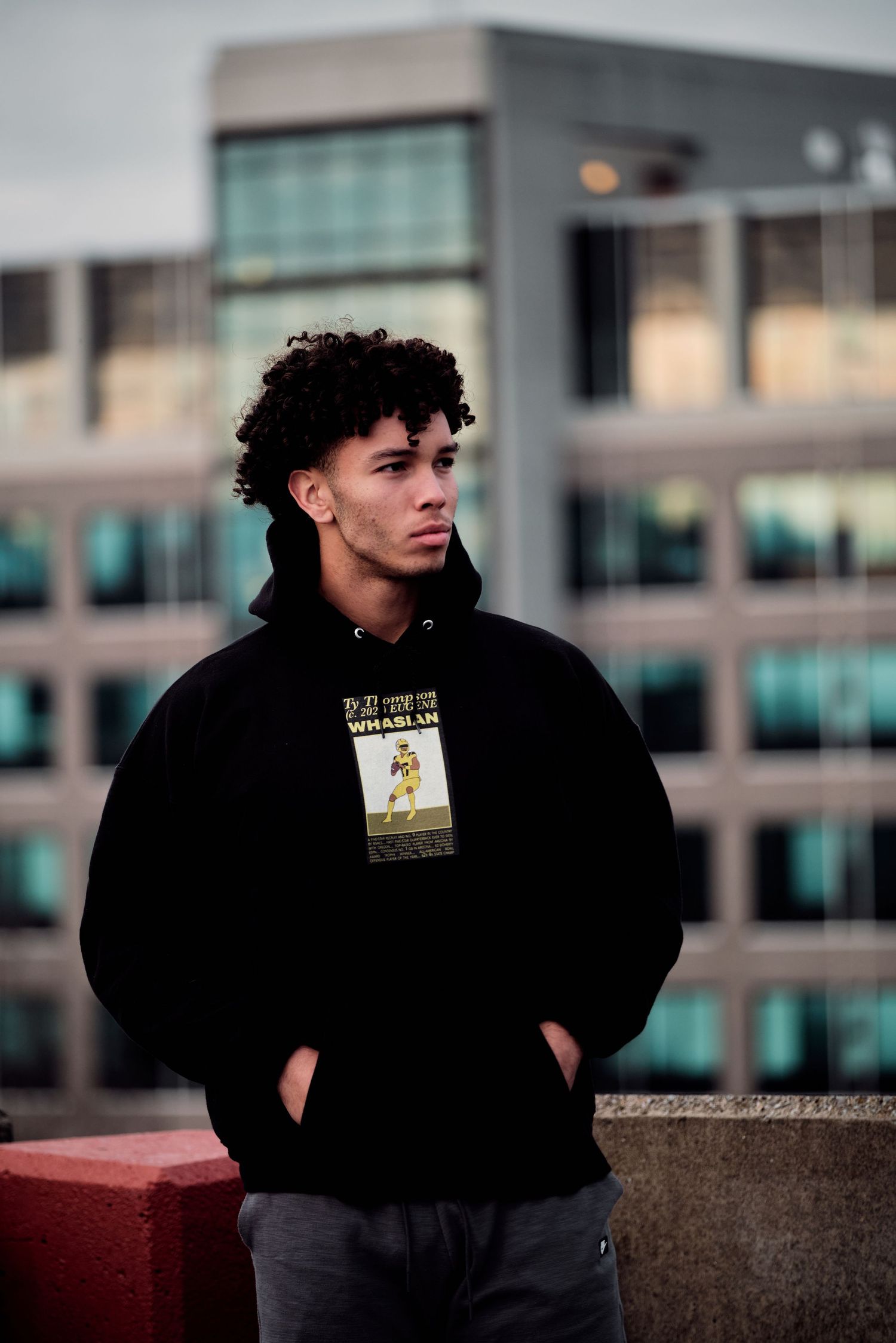 Person in black hoodie poses near urban buildings at dusk.