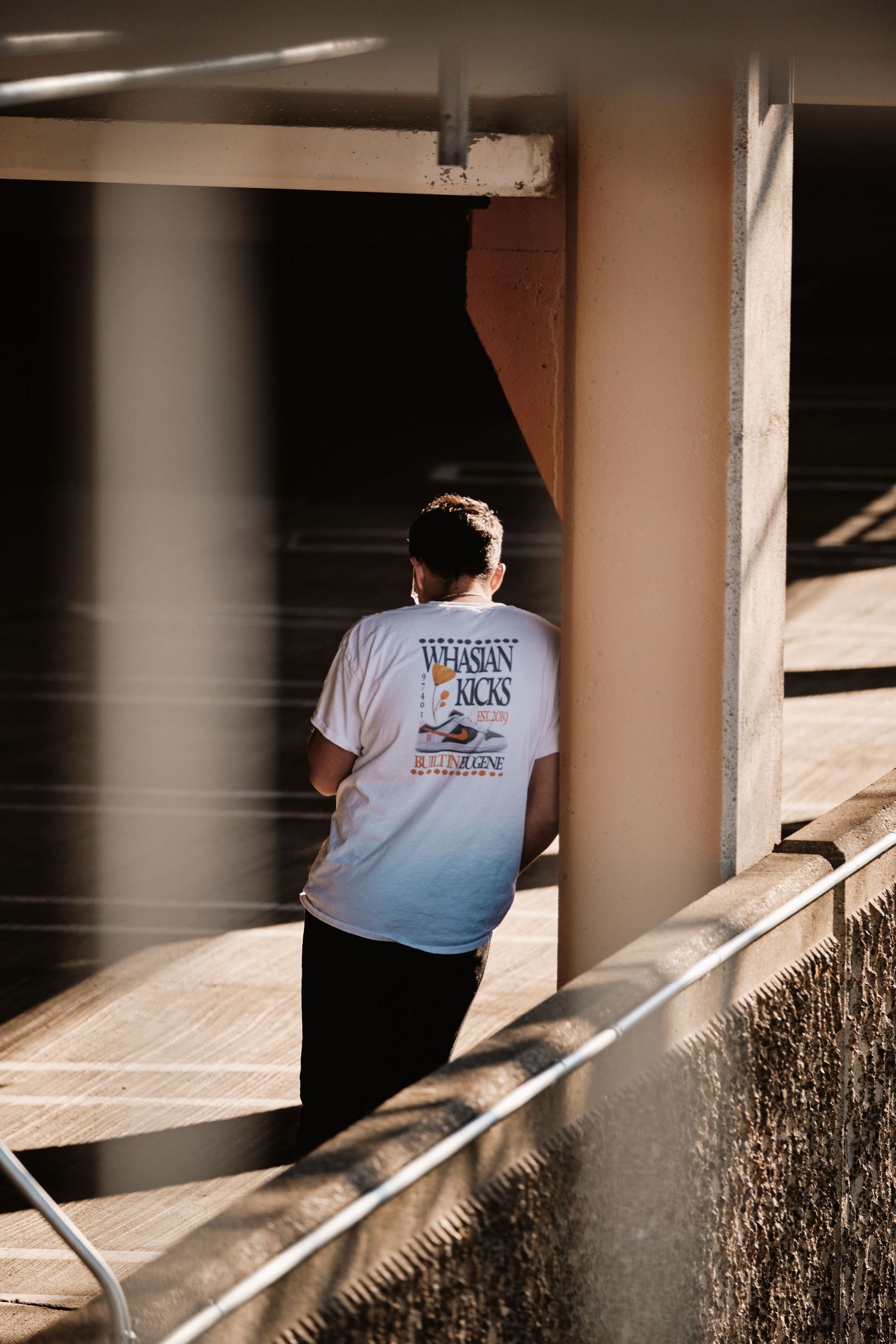Person in white t-shirt leans on concrete pillar in shadow.