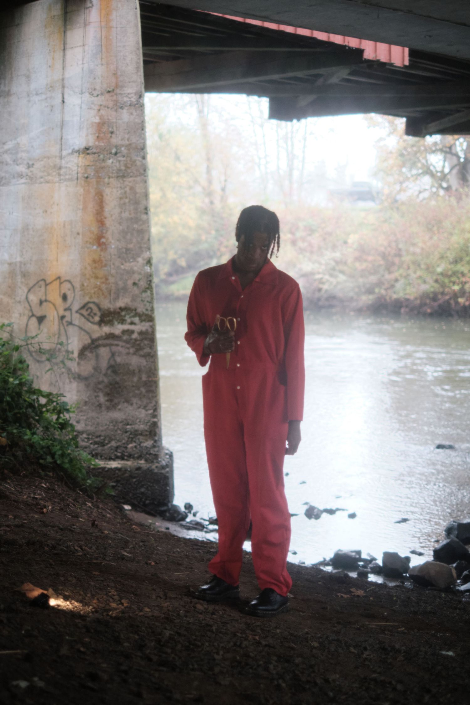 Figure in red stands under a bridge near a river on an overcast day.