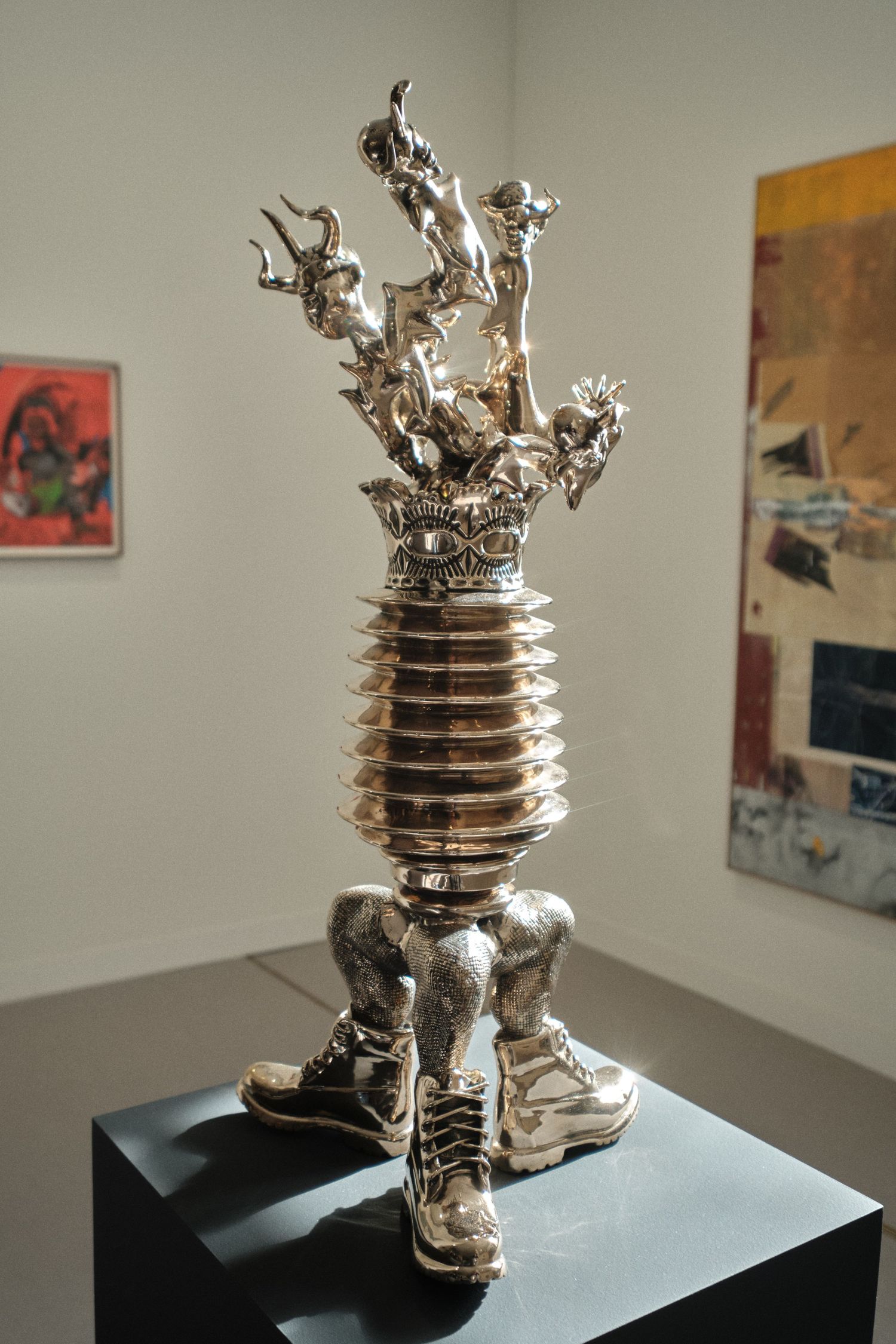 Metallic sculptural artwork with crown-like top displayed in art gallery setting.