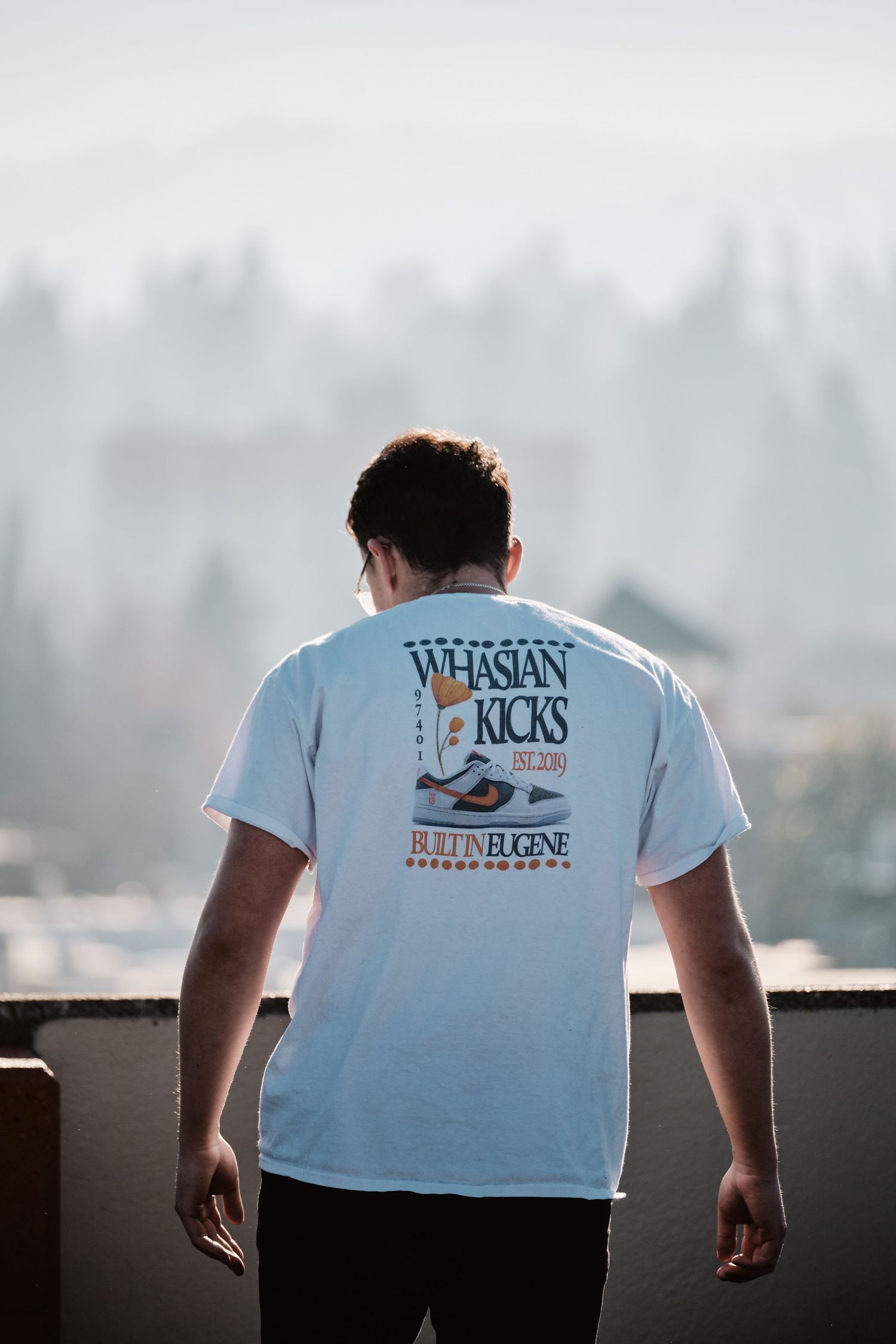 Person wearing a white t-shirt with kicks branding stands against a misty background.