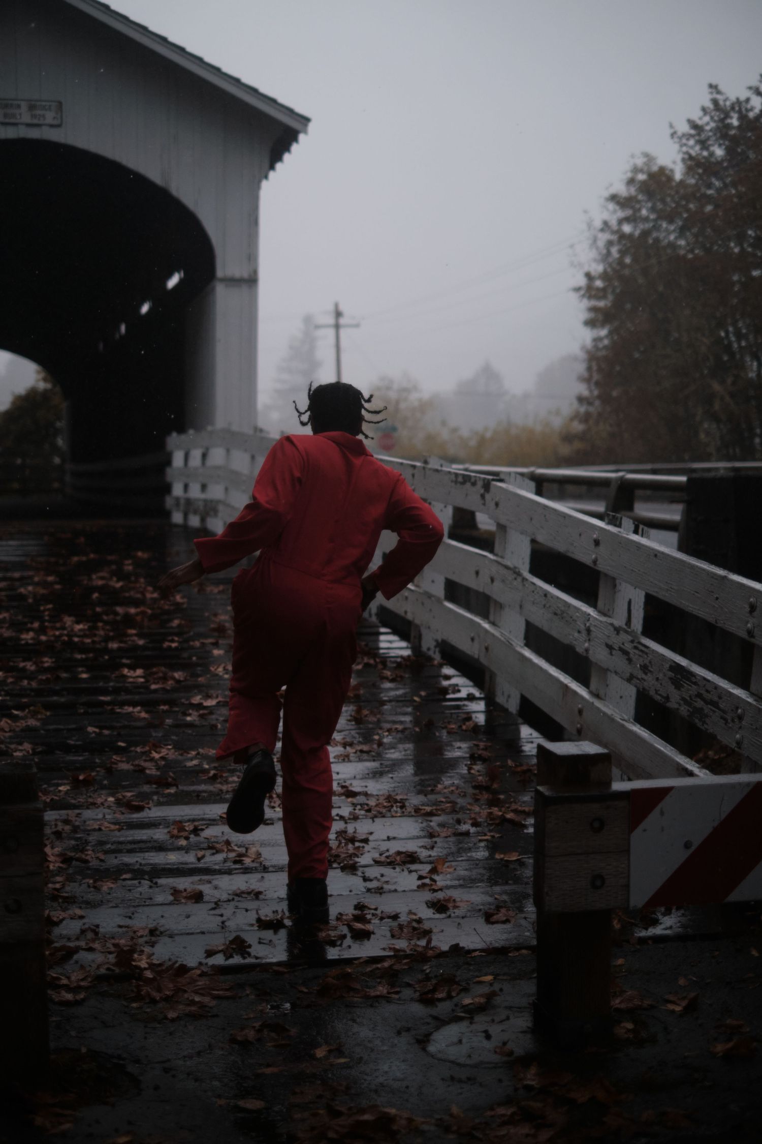 Figure in red hopping along through a misty covered bridge strewn with autumn leaves.