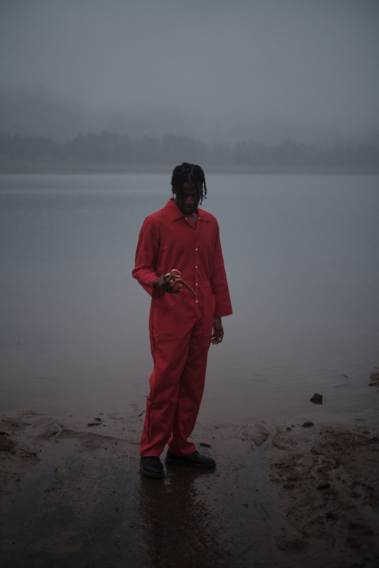 Mysterious figure in red jumpsuit stands in shallow water during overcast day.