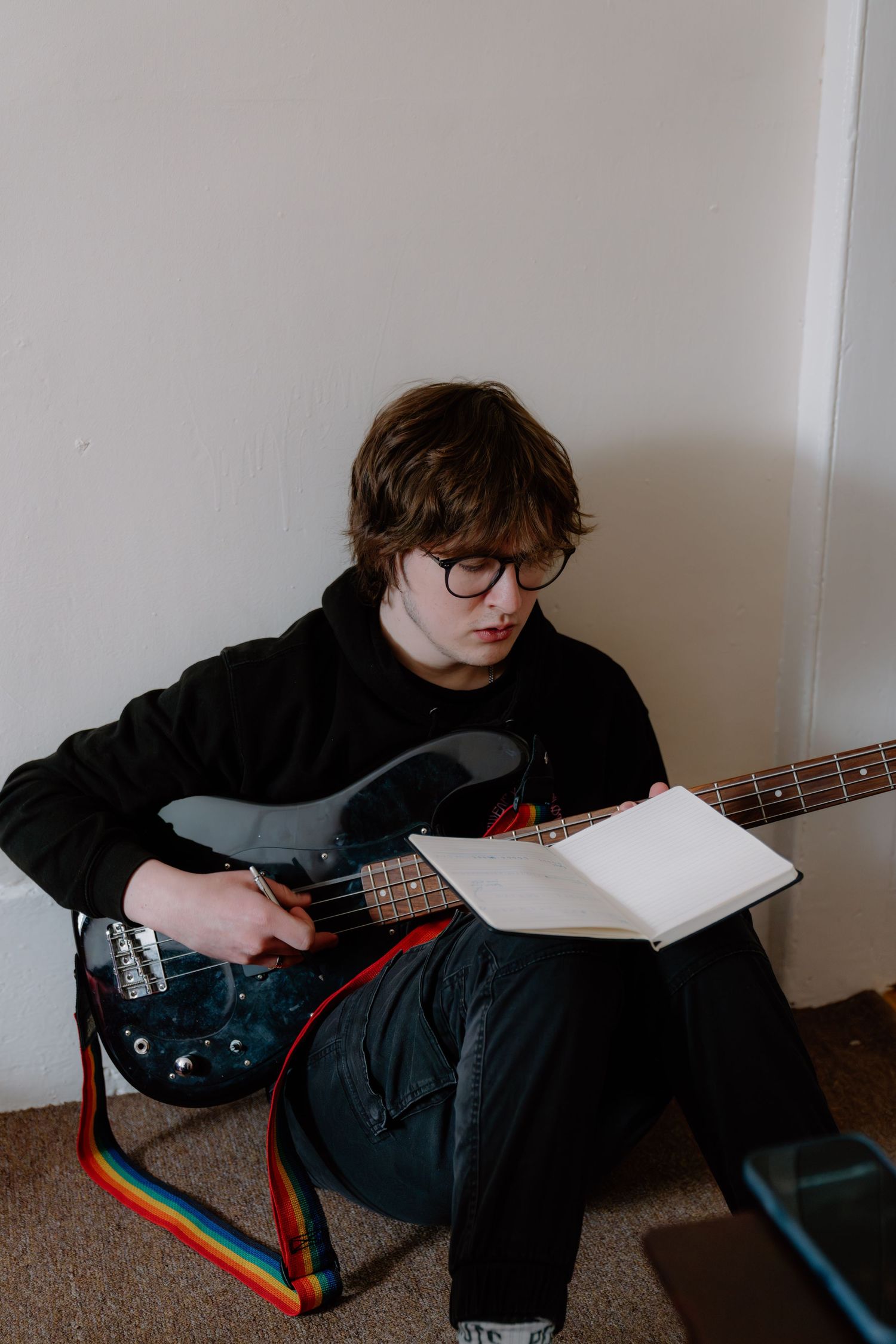 Teen with glasses sits on floor writing in notebook while holding a black electric guitar with rainbow strap.