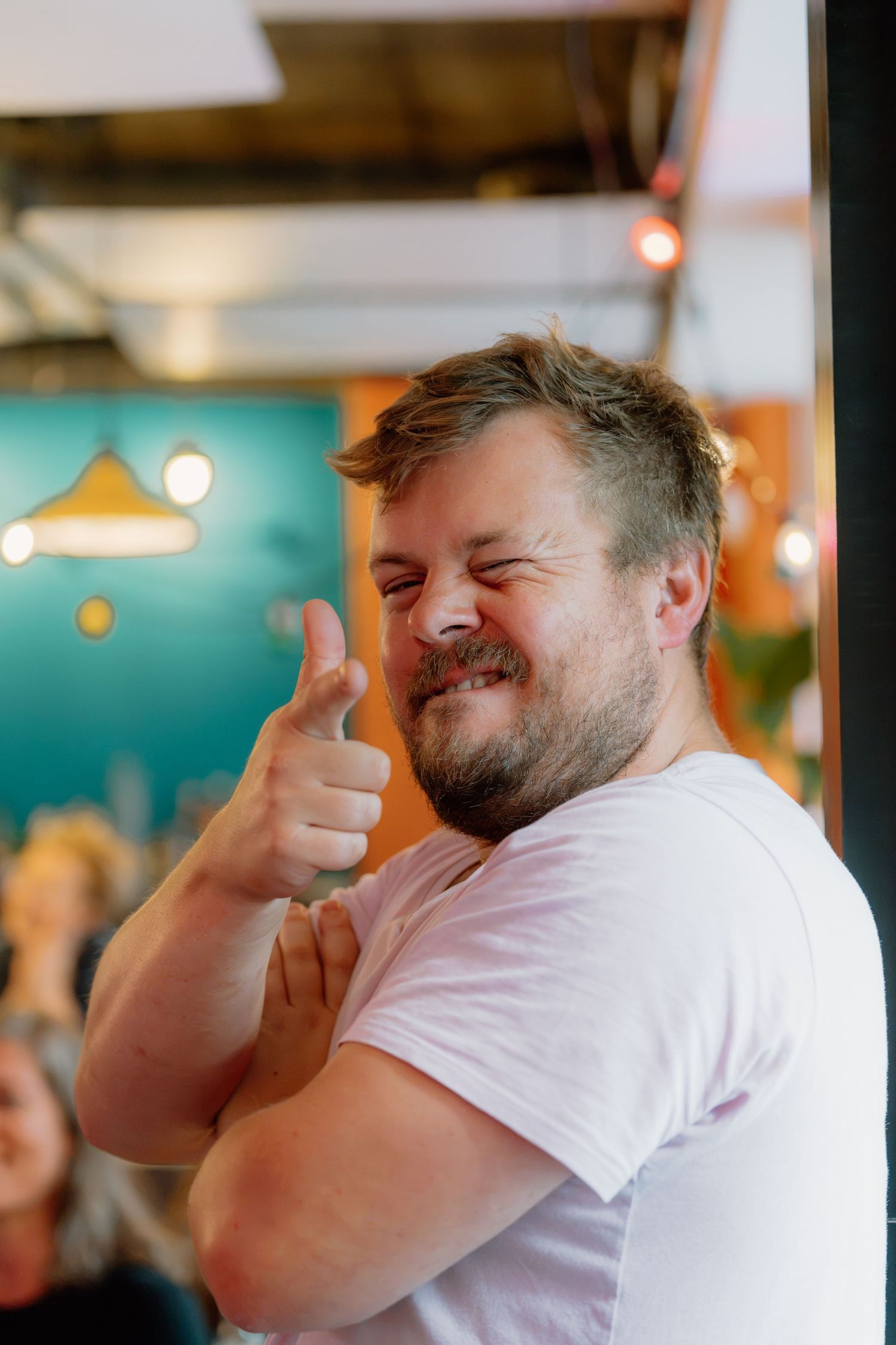A cheerful bearded man in a white t-shirt gives a thumbs up and winks in a colorful cafe setting.