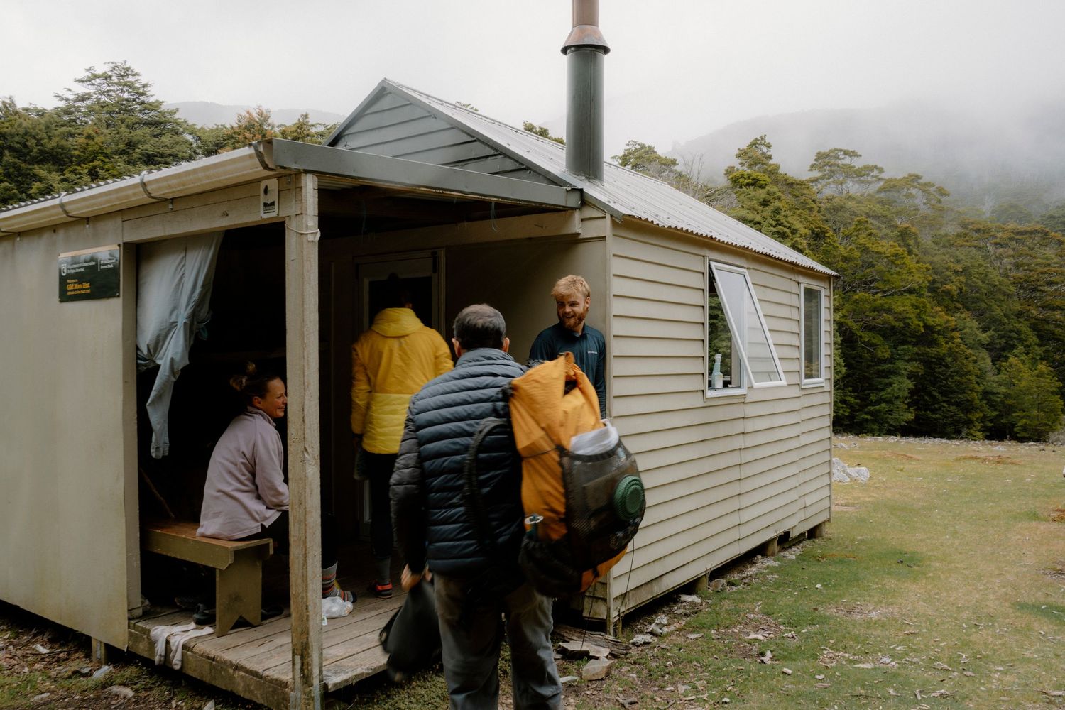 A group of hikers enter a rustic white mountain hut shelter on a misty hillside trail.