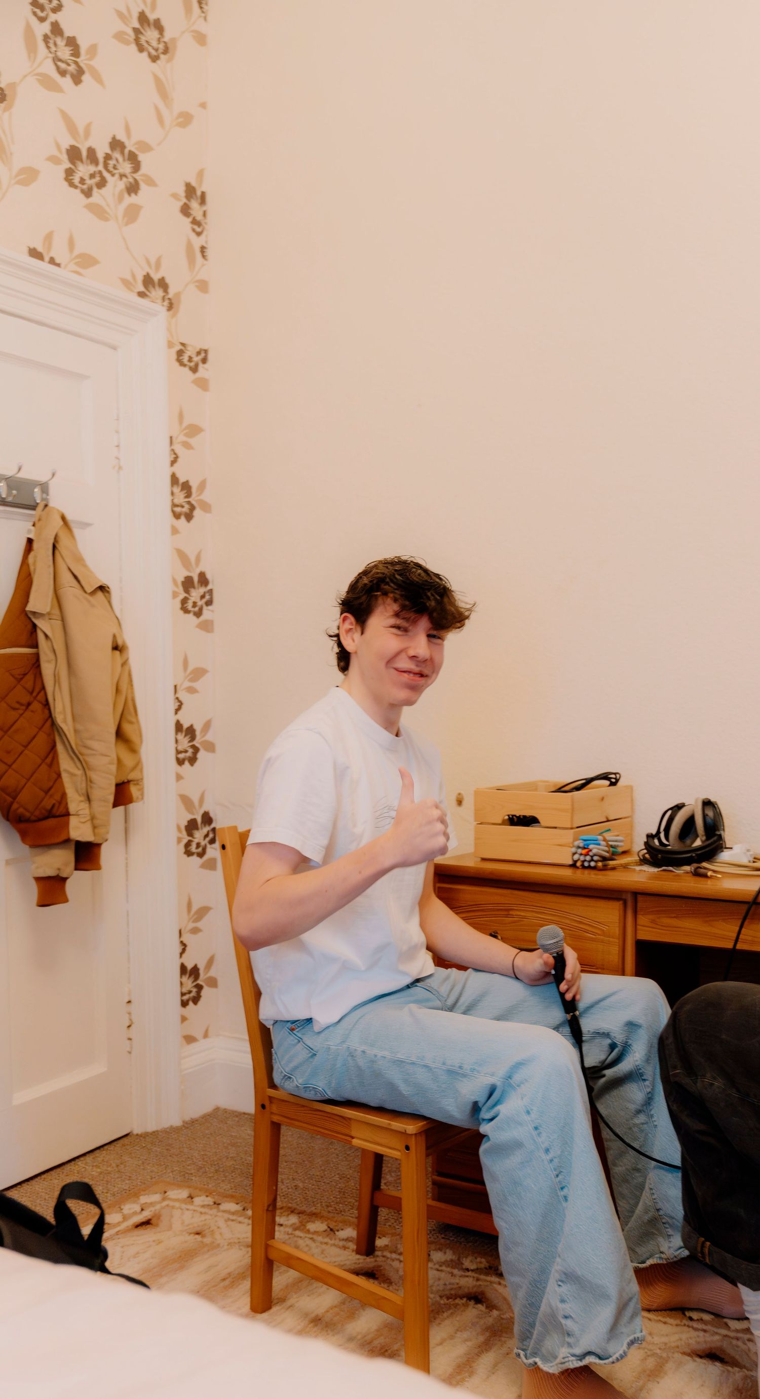 Young man in white t-shirt and jeans sits on wooden chair giving thumbs up in a cozy room with floral wallpaper.