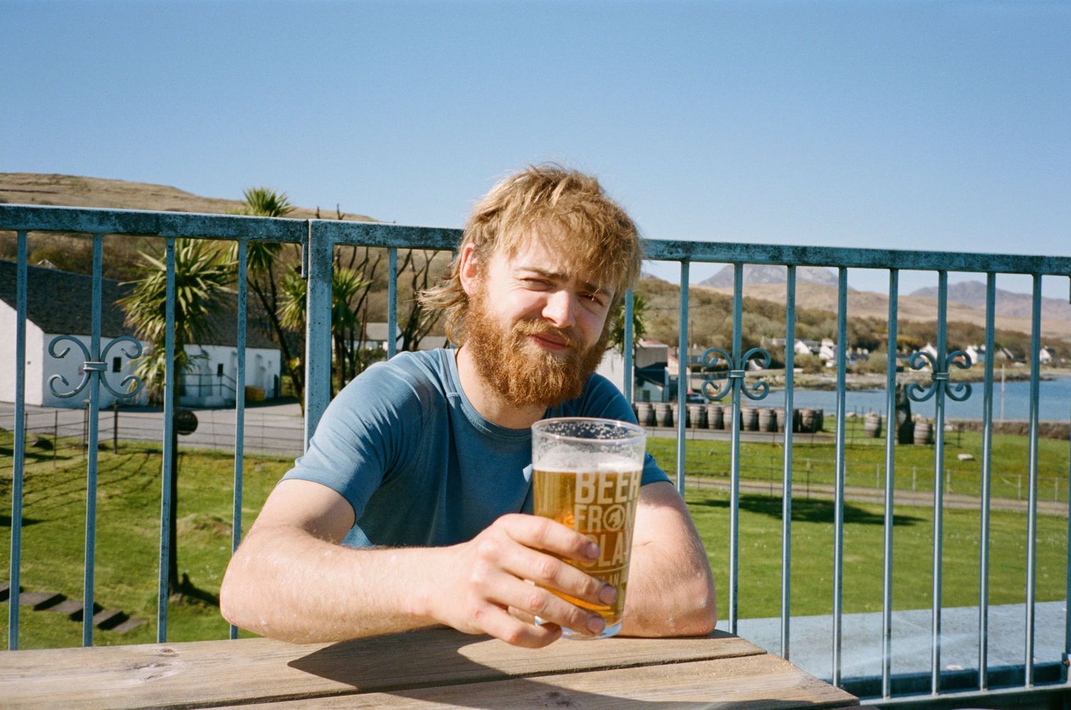 Smiling bearded man holding a pint of beer at an outdoor table with scenic coastal views on a sunny day.