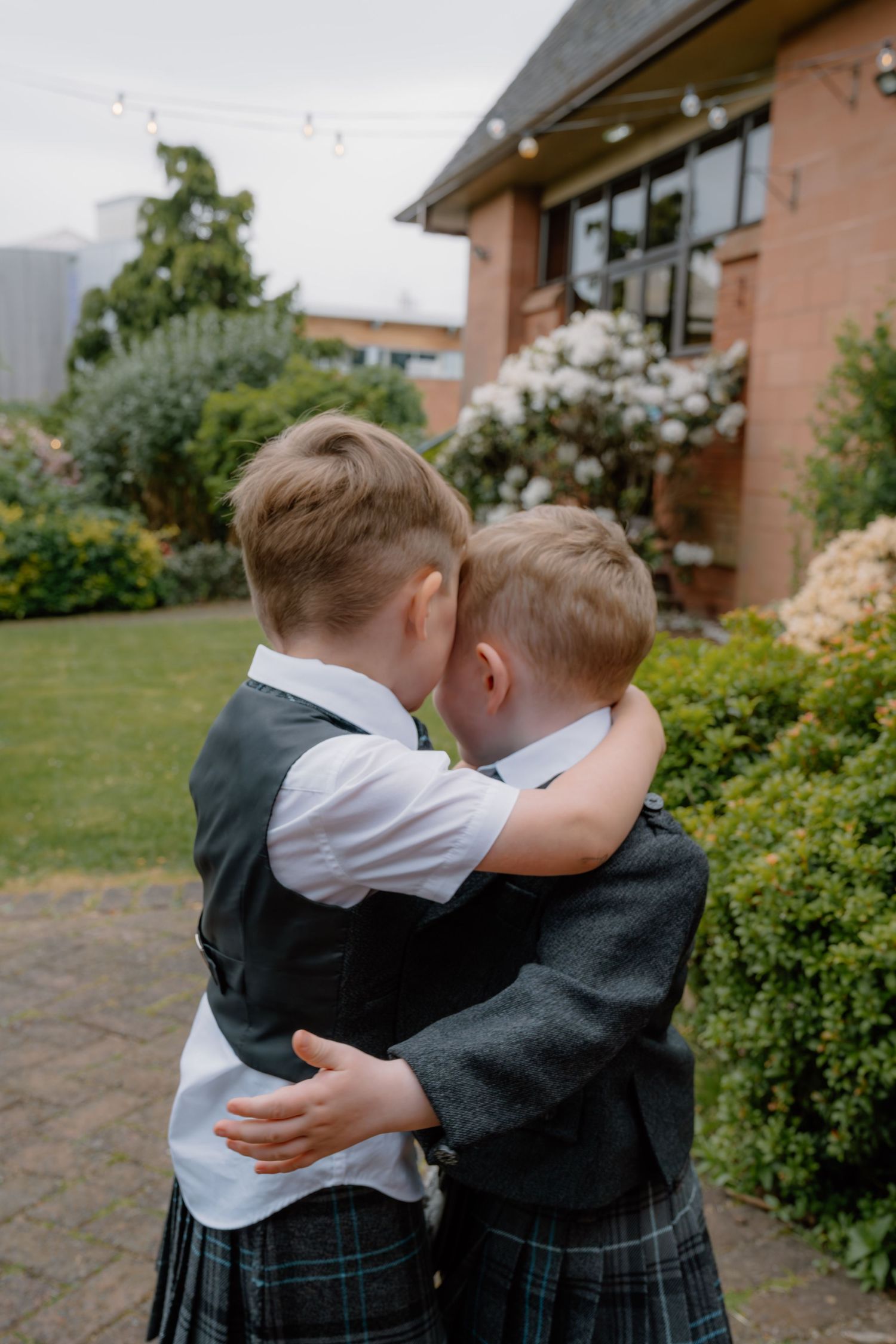 Two young boys in kilts and waistcoats share a sweet hug in a garden setting at a wedding.