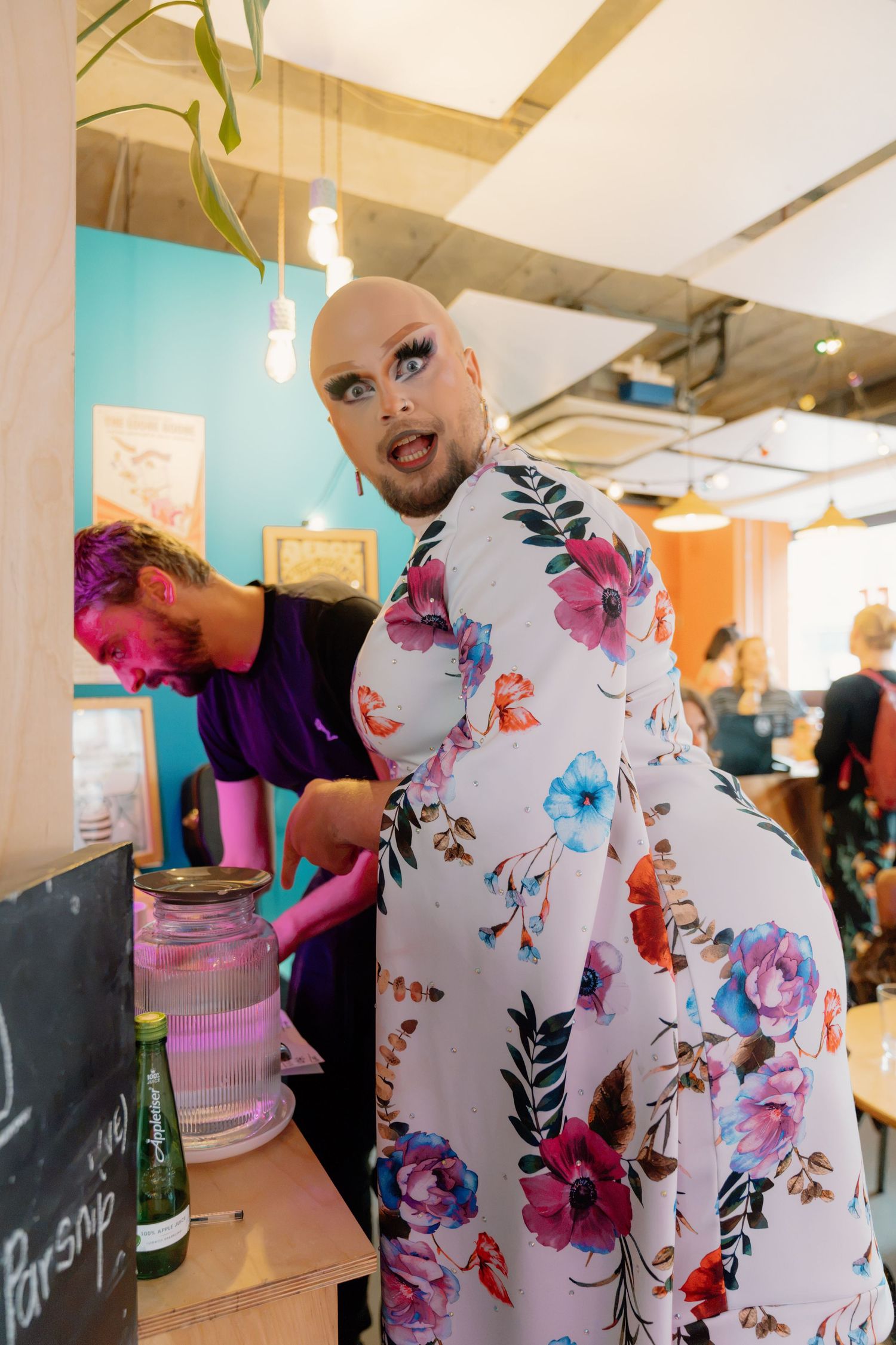 Bald drag queen in floral jumpsuit with dramatic eye makeup poses at a colorful cafe counter.