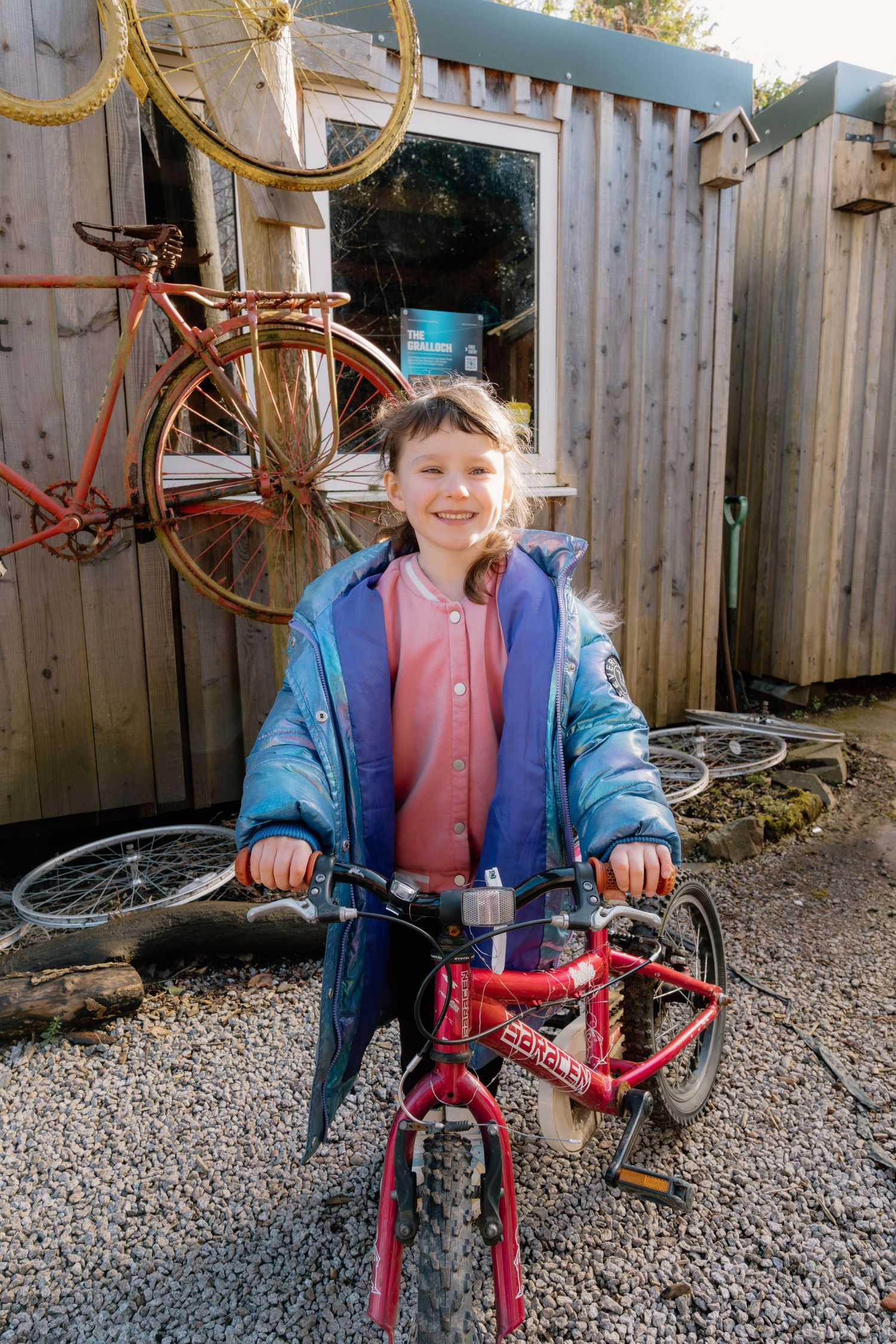 Smiling girl in blue jacket riding a red children's bicycle outside a wooden shed with vintage bikes.
