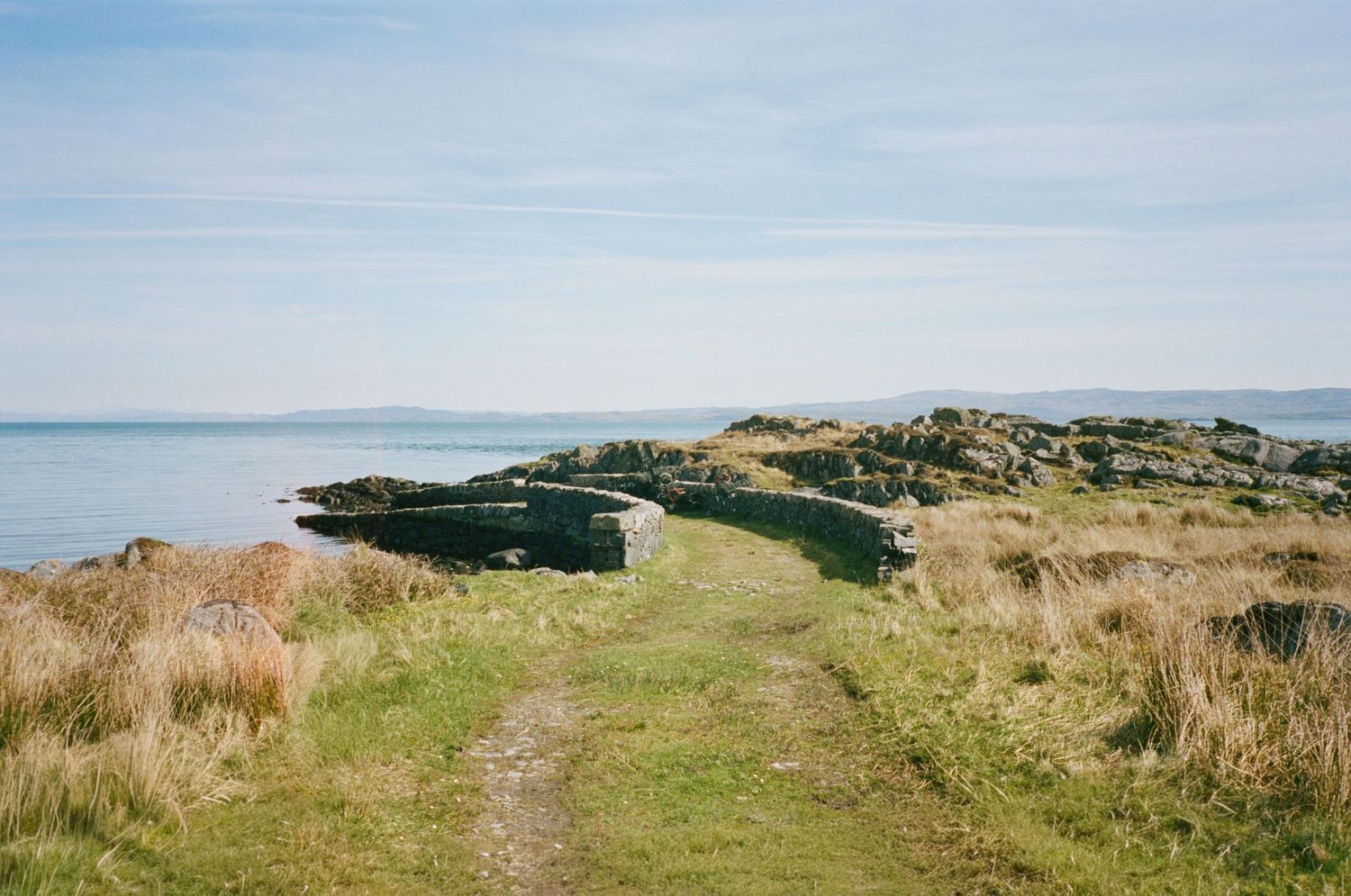 Scenic coastal path leading to rocky shoreline with wild grass, calm sea, and blue sky in background.