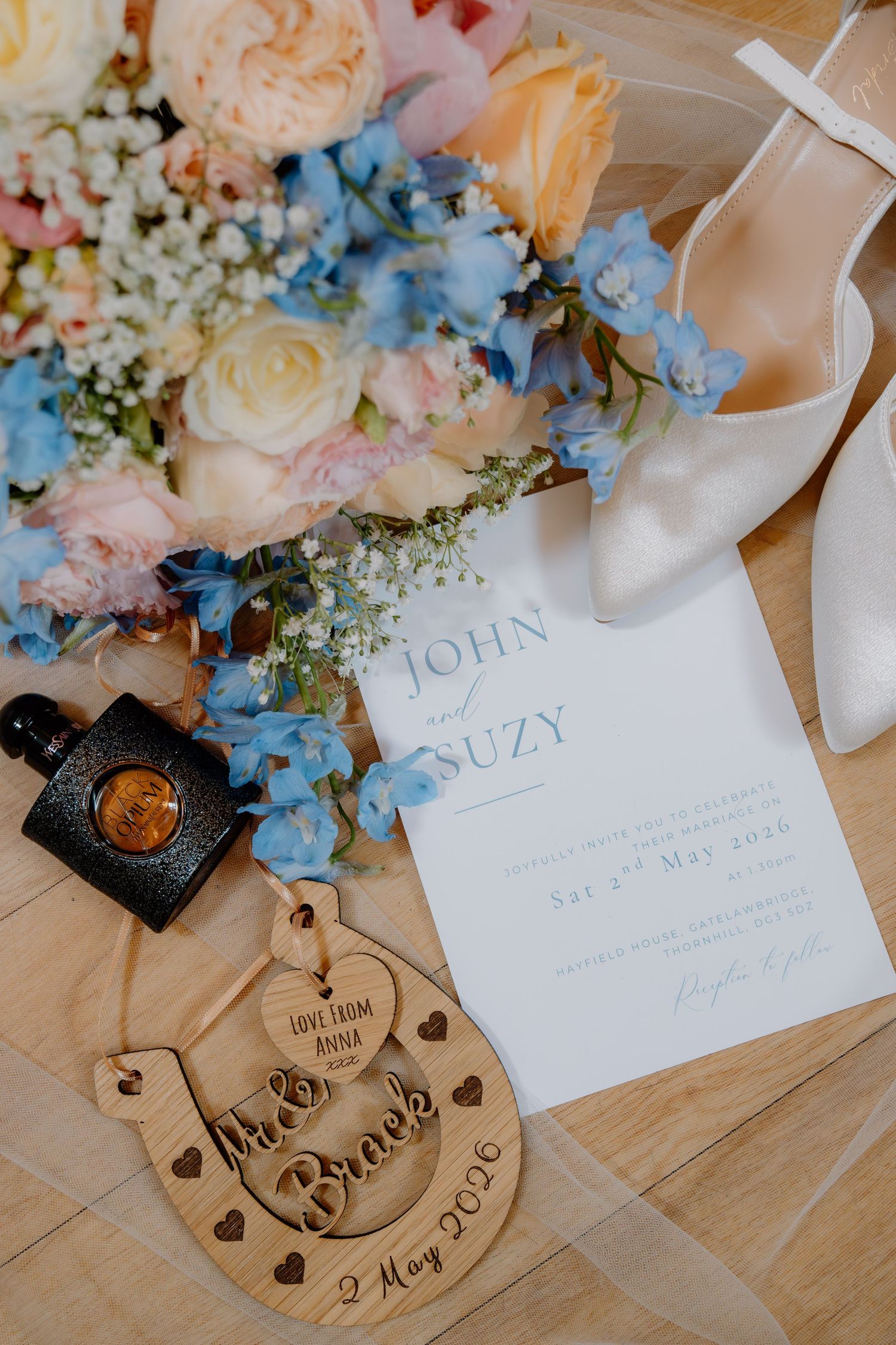 Elegant wedding flat lay with floral bouquet, white heels, perfume, wooden keepsake, and invitation for John and Suzy.
