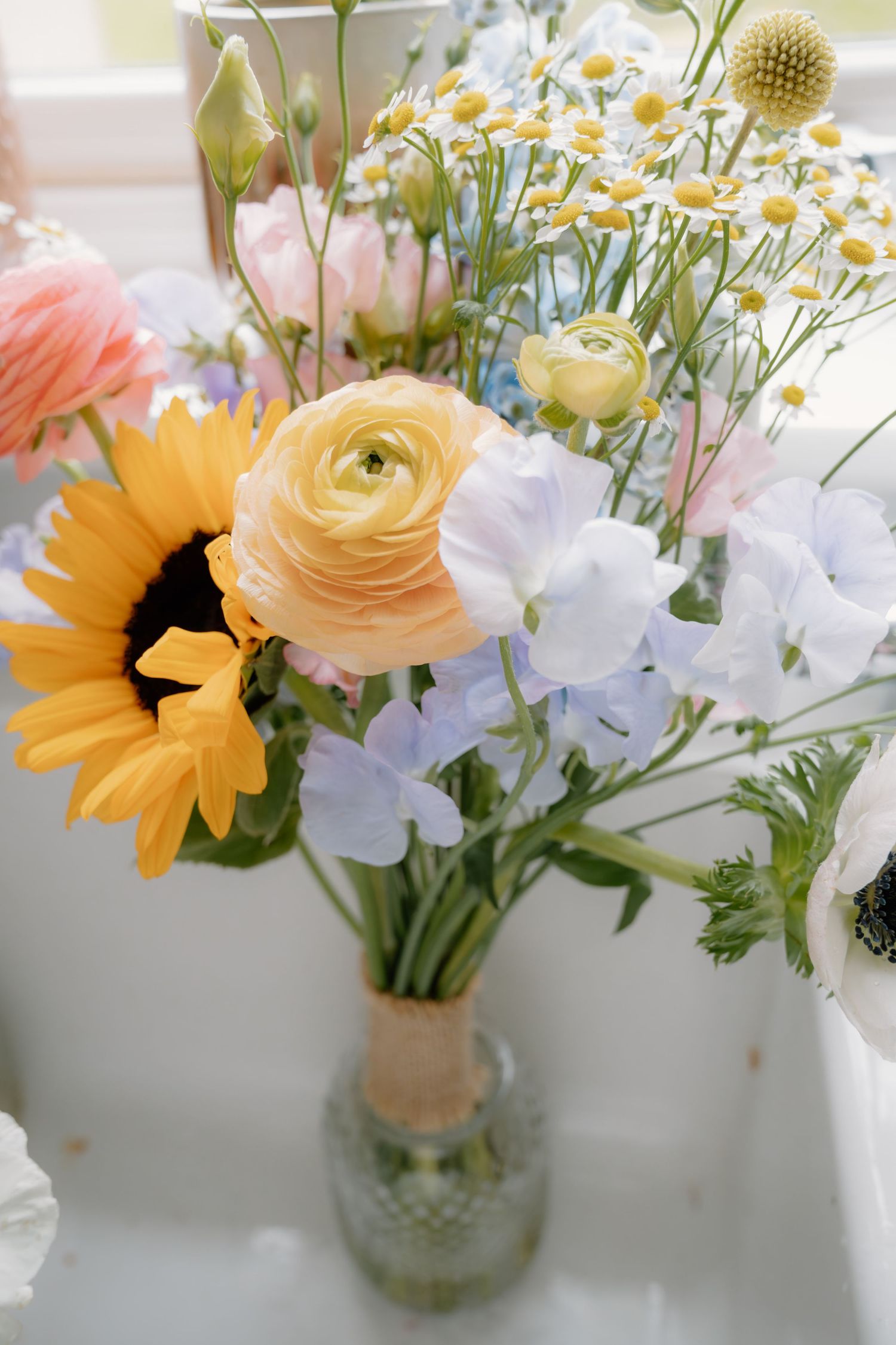 A beautiful wedding bouquet featuring sunflowers, ranunculus, sweet peas, and daisies in pastel hues arranged in a glass vase.